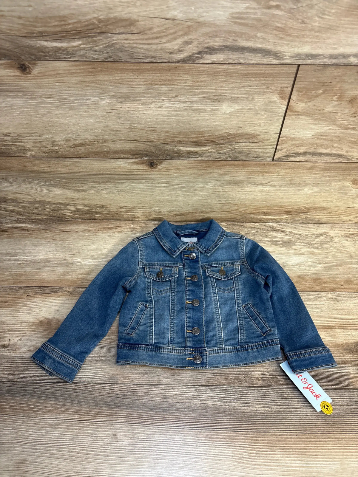 Kids denim jacket with button front and chest pockets, laid flat on wood floor, brand tag visible