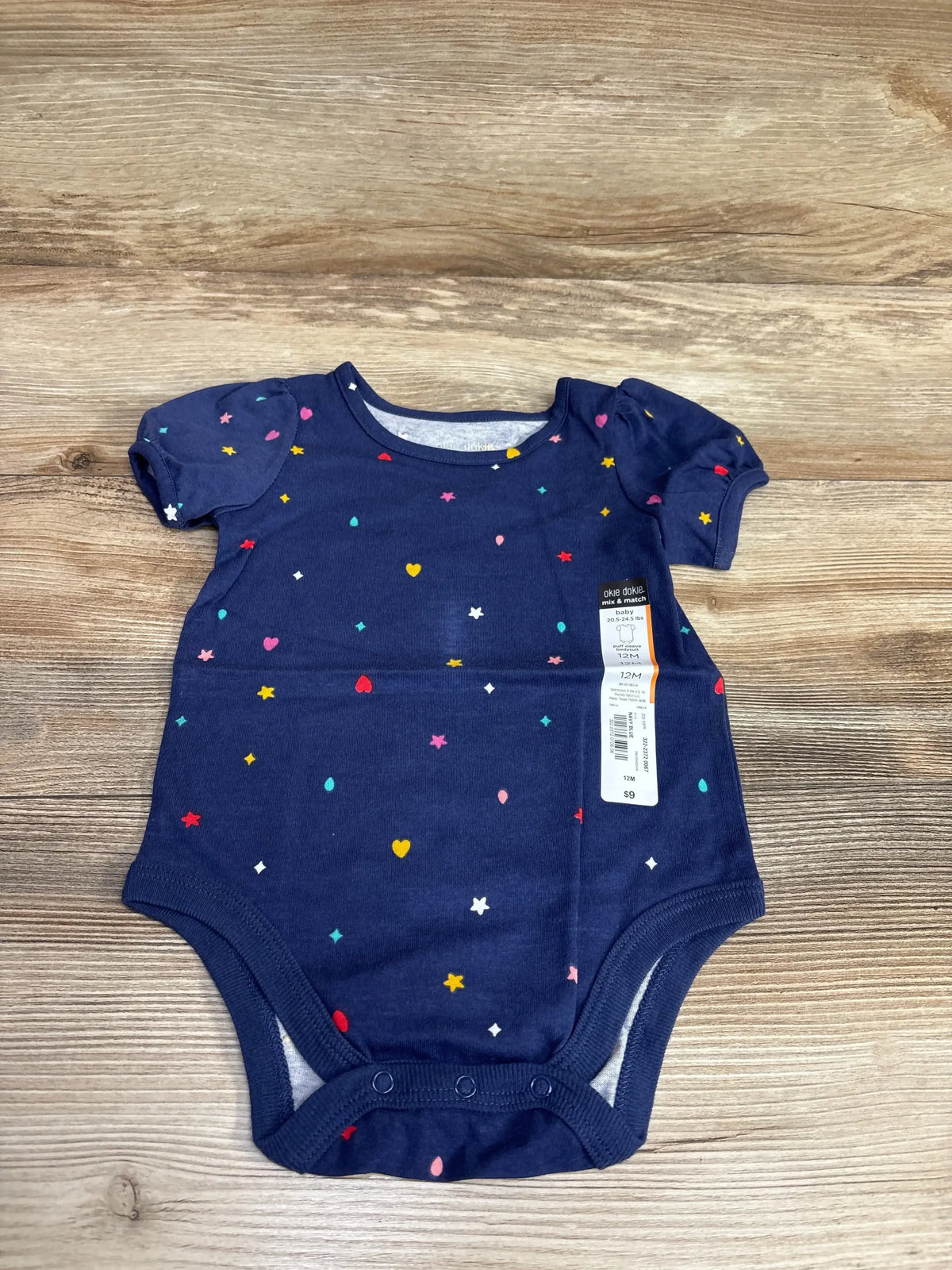 Top-down view of Okie Dokie navy puff-sleeve bodysuit with multicolored hearts and stars and visible 12M tag on wood