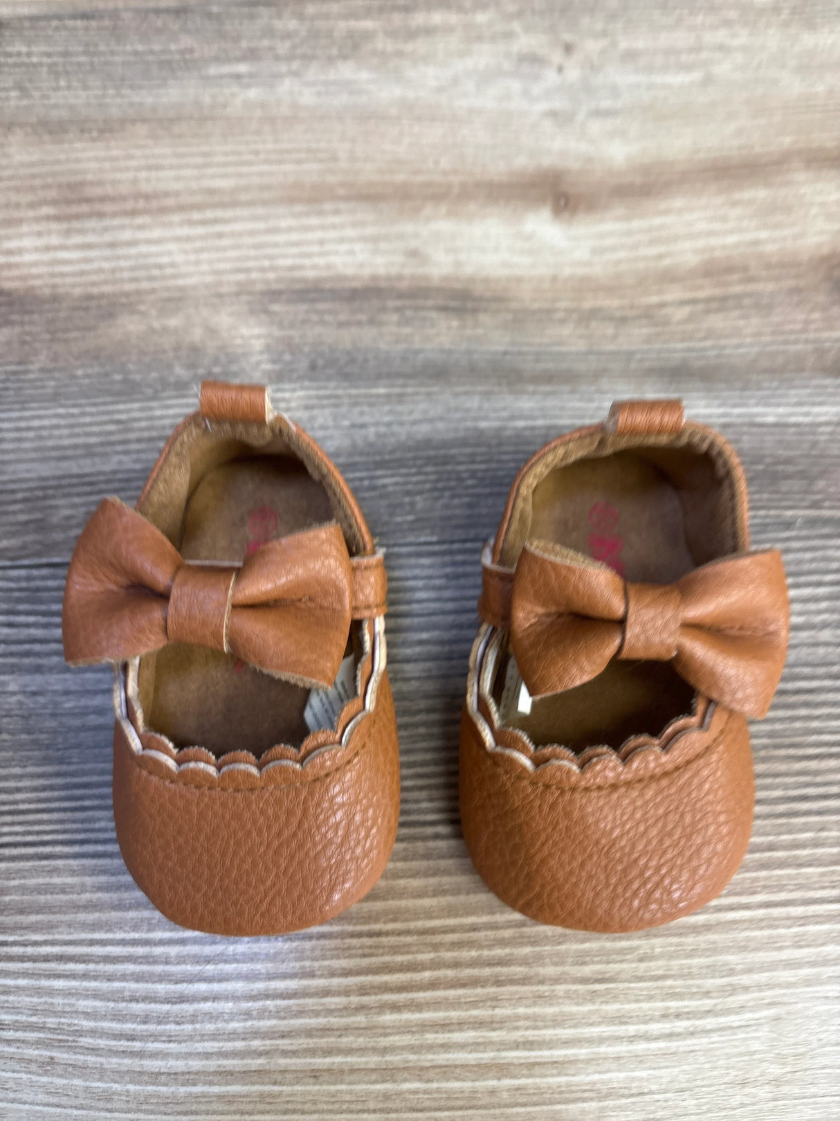 Top-down view of Nuby anti-slip first walking shoes in brown size 2C showcasing leather bow and scalloped edge with wooden background