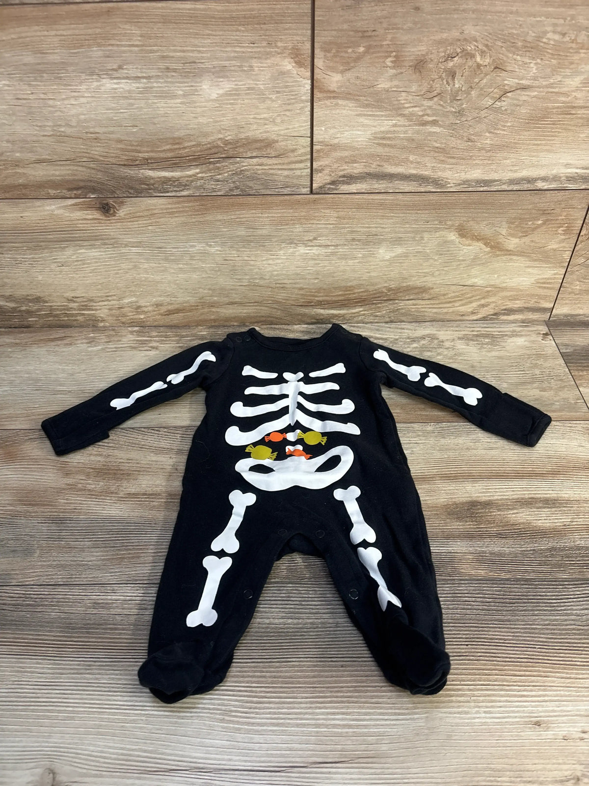 Front view of Just One You black skeleton footed coverall showcasing white skeleton print and covered feet on a wood-look floor