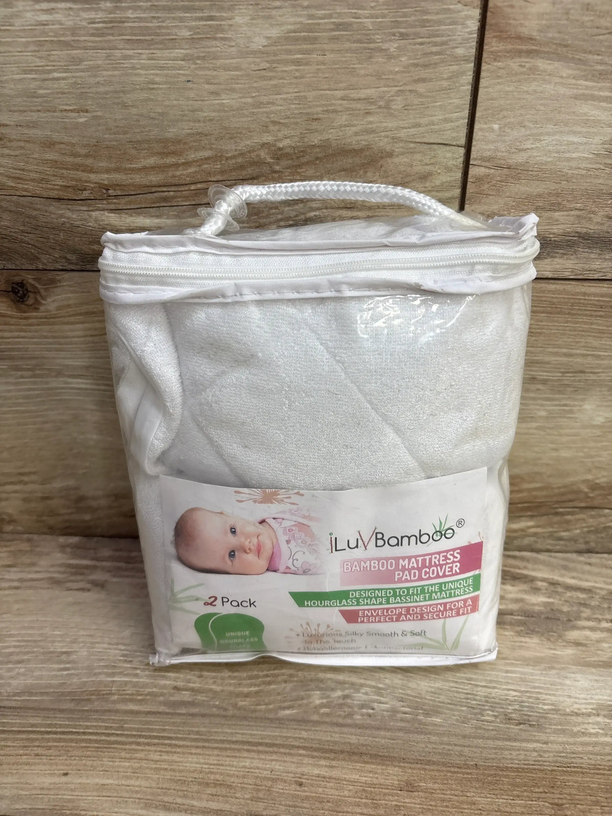 Front view of ILuvBamboo 2-pack white bamboo bassinet mattress pad covers in clear zip pouch showing product label on wood background