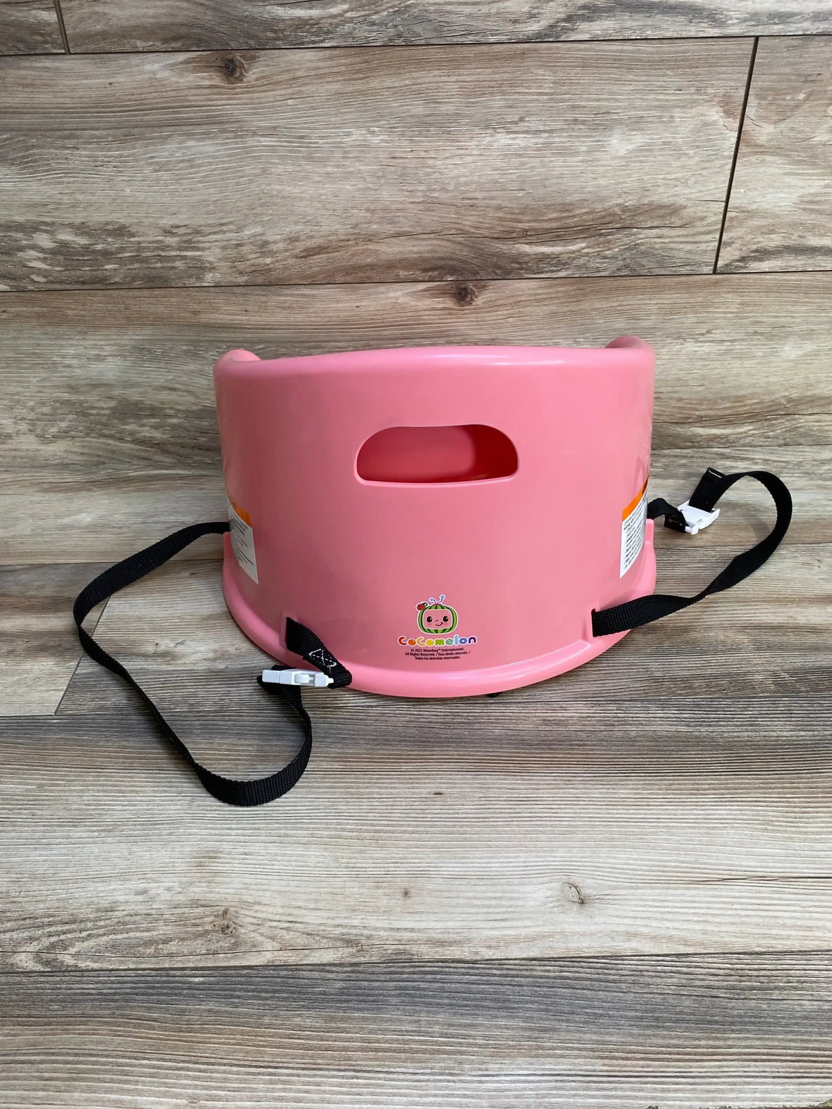 Back view of Cocomelon 15-inch pink booster floor seat showing carry handle and black safety straps on wood floor