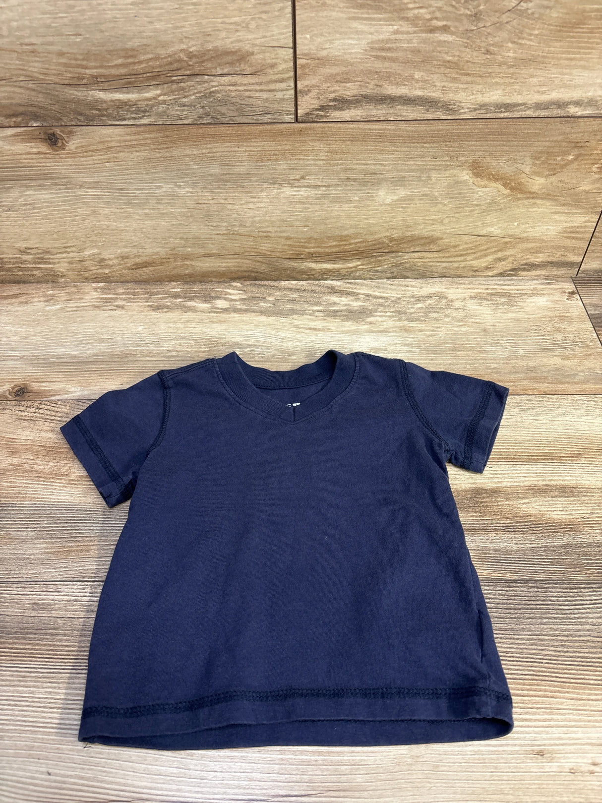 Flat lay of navy toddler V-neck T-shirt, size 2T, showing short sleeves and visible hem stitching on wood floor background