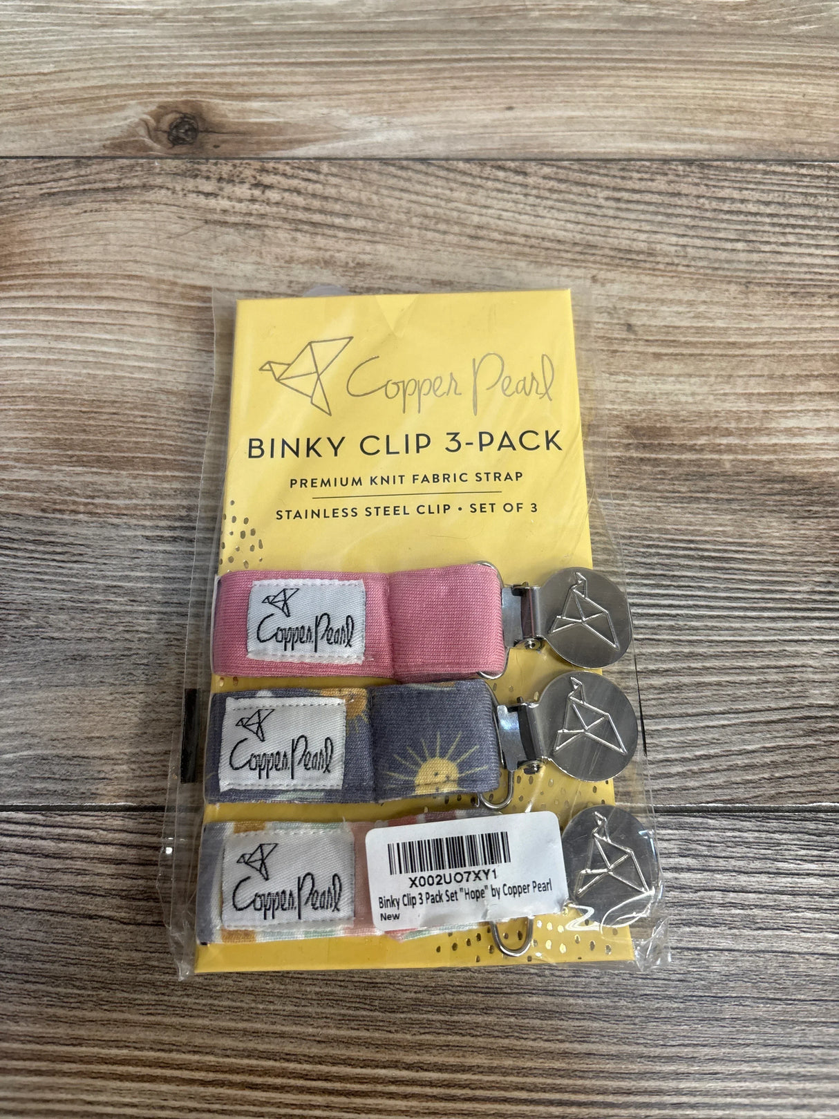 Top-down Copper Pearl binky clip 3-pack with three knit straps (pink, gray sun, peach) and metal clips on wood background