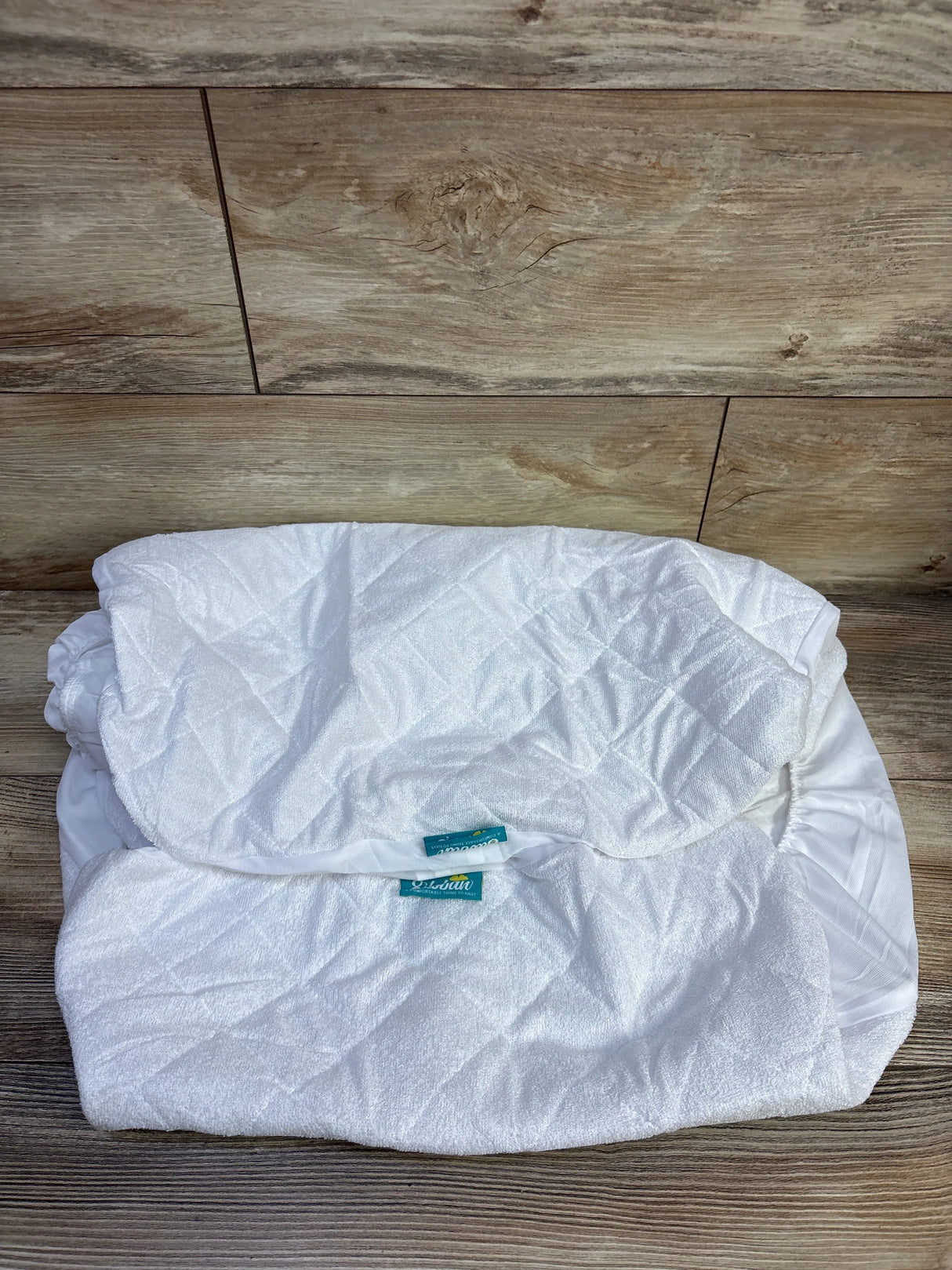Top-down view of folded white quilted waterproof bassinet mattress pad cover showcasing elastic skirt and diamond quilting on wood floor background