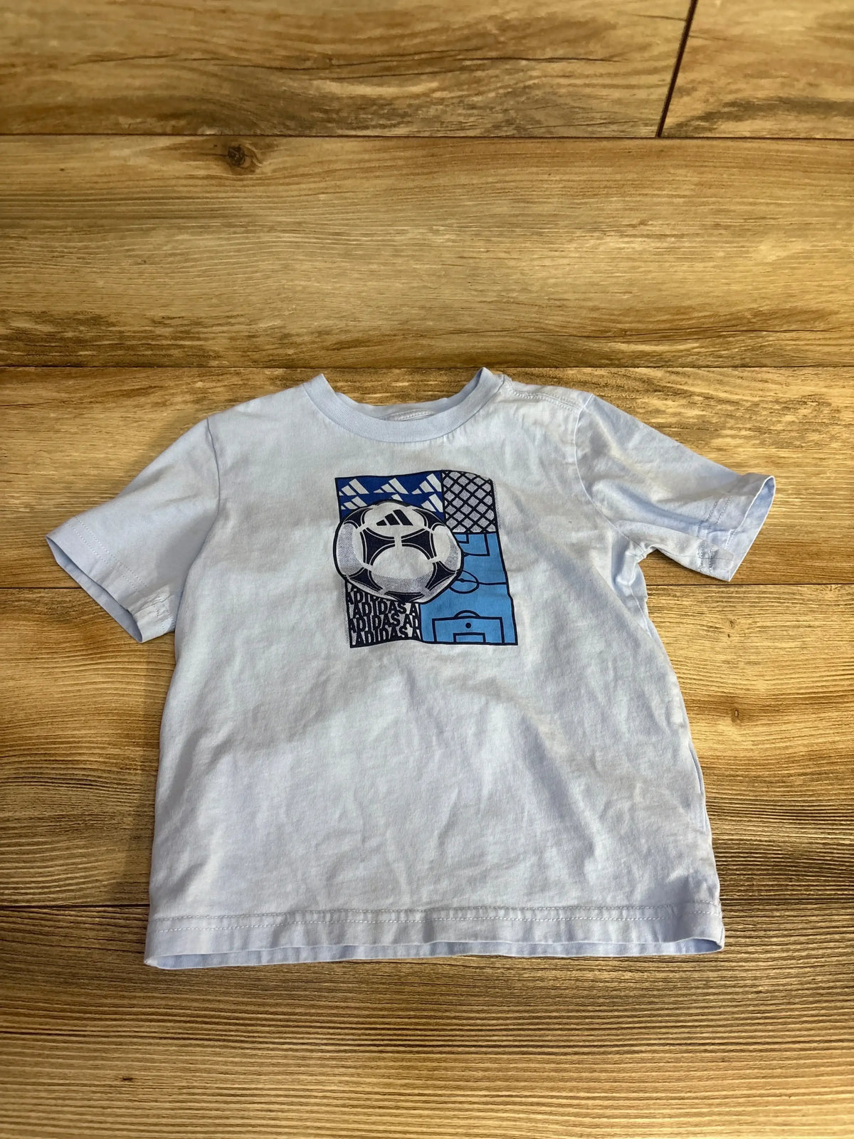 Light blue toddler T-shirt with a soccer ball and goal graphic design on wooden floor