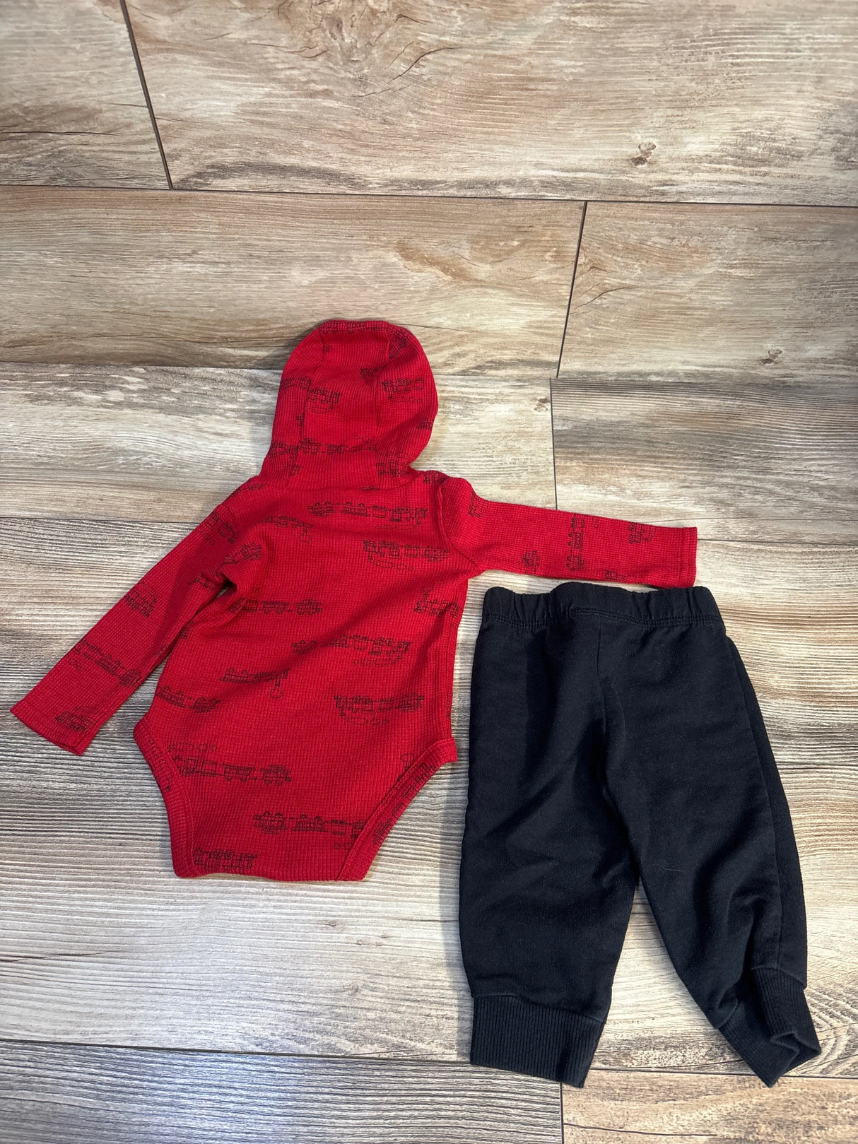 Red baby hooded bodysuit with train print and black pants set on wooden floor
