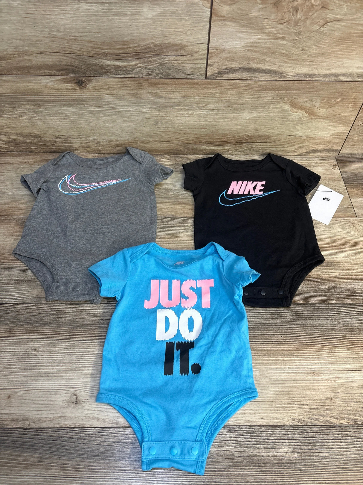 Three Nike baby bodysuits in gray, black, and blue with slogans, displayed on wooden floor.