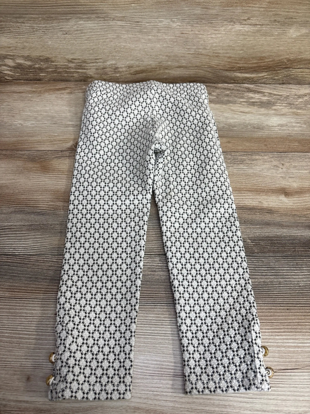 Flat lay of girls' Janie & Jack white ponte pants (3T) showing black geometric pattern and gold button cuffs on wood background
