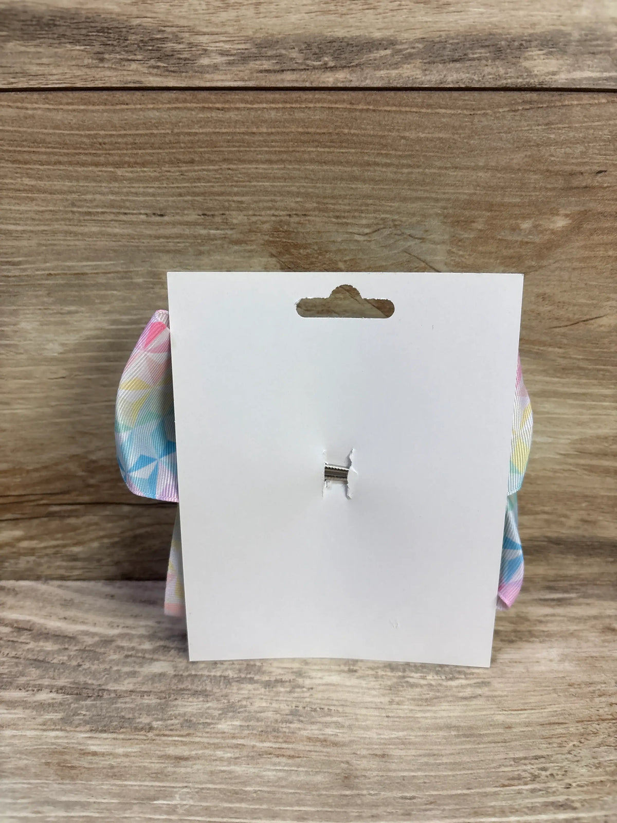 Back view of large geometric hair bow showcasing pastel multicolor pattern and metal clip on a white display card over a wood surface