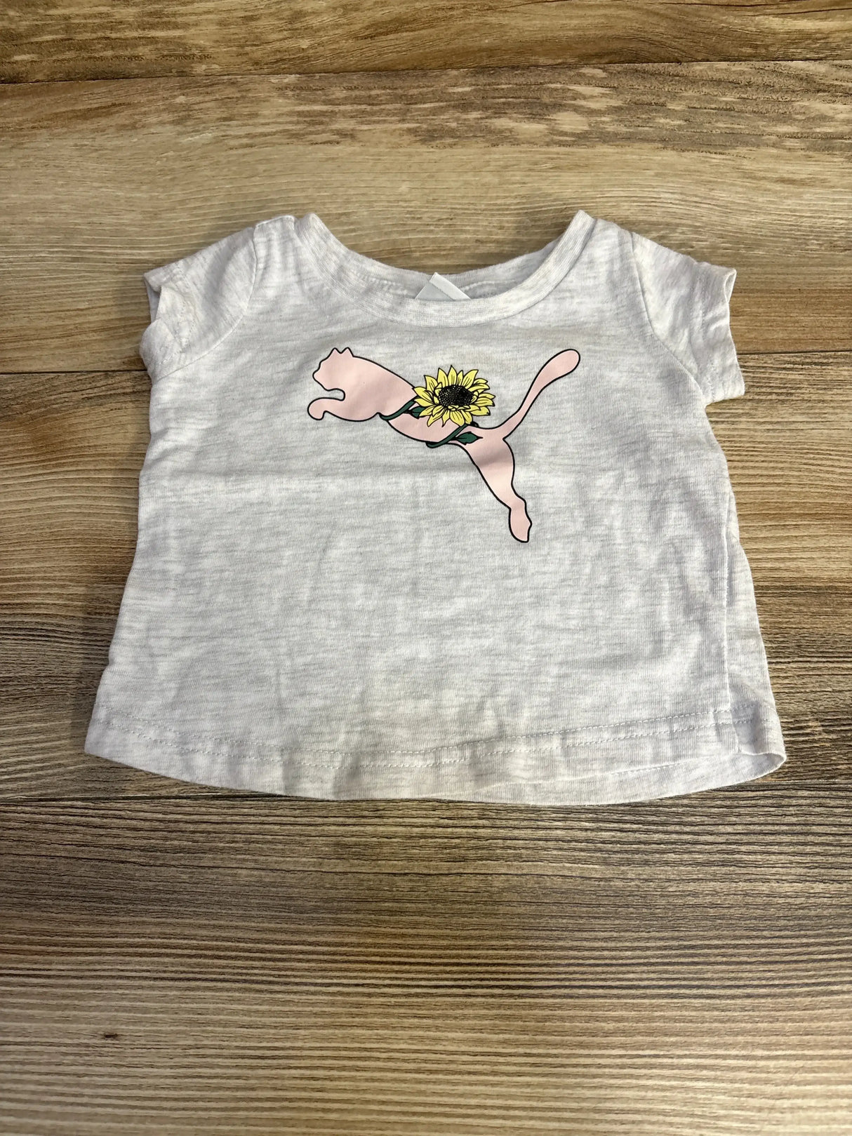 Front view of grey baby Puma shirt with pink leaping-cat logo and sunflower on chest on wood background