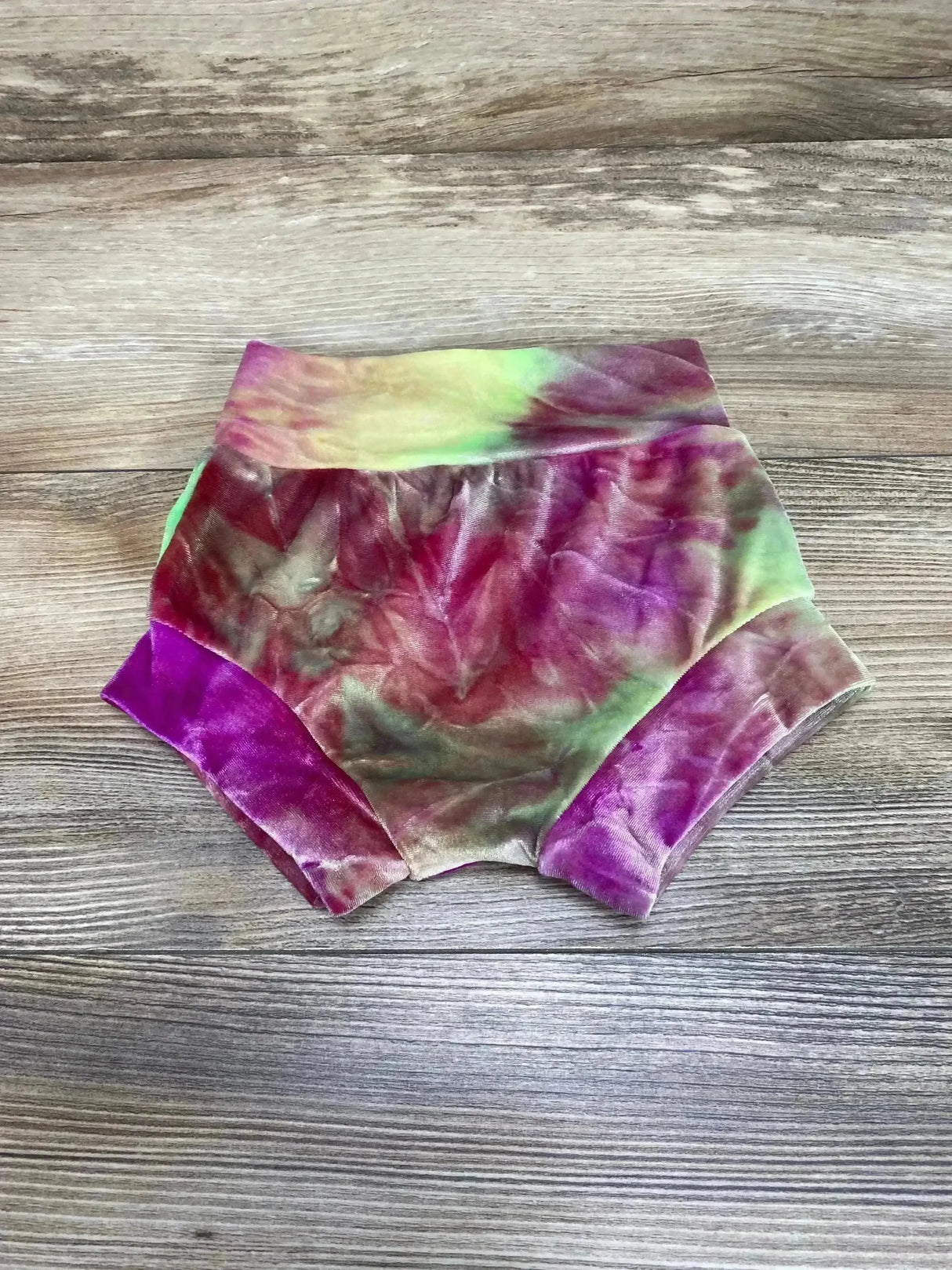 Top-down view of velvet baby bloomers showcasing green and purple tie dye fabric and wide elastic waistband on wood floor