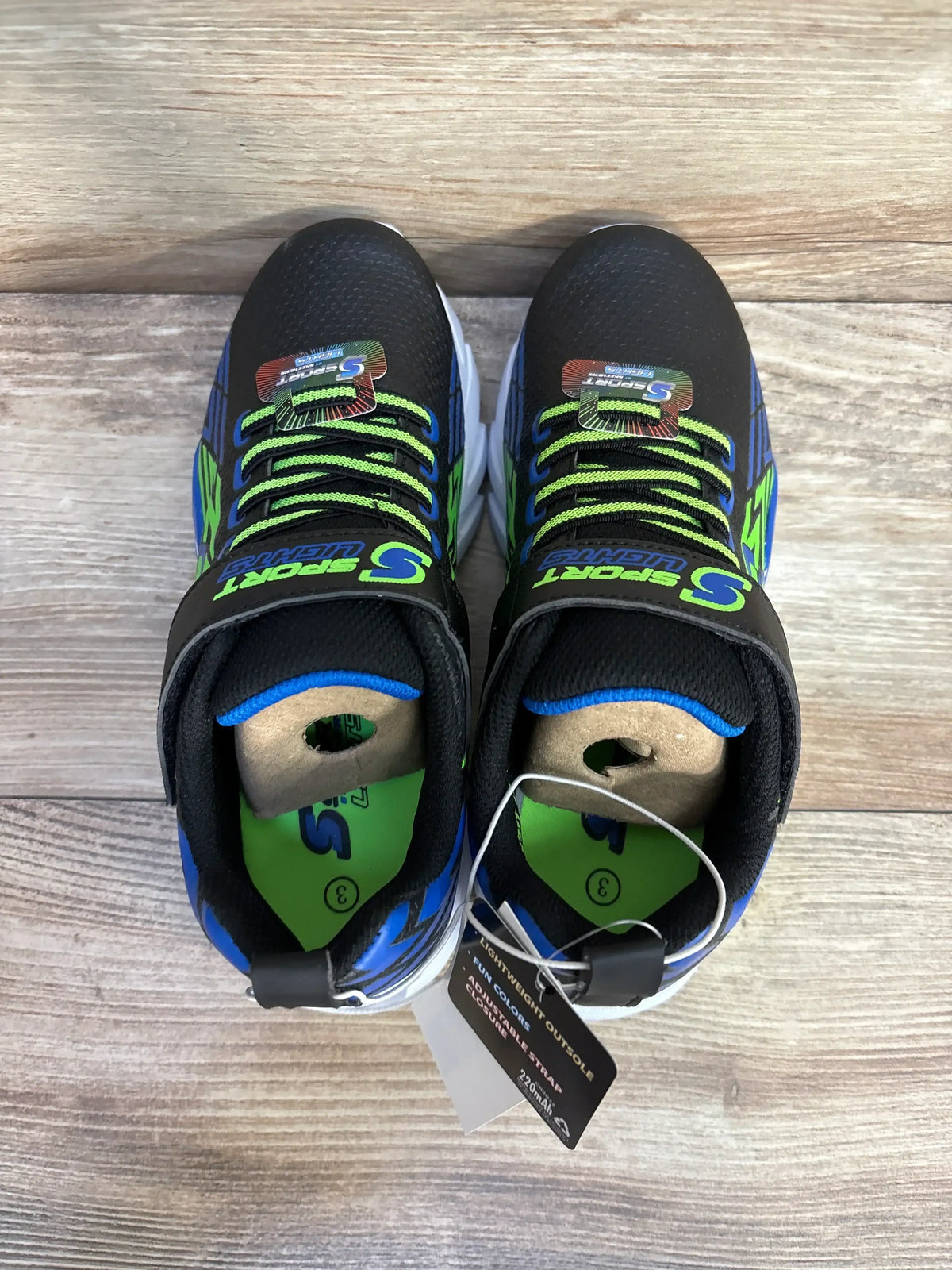 Top view of S Sport by Skechers Lights Ottis sneakers showcasing black mesh upper with green and blue accents and hangtag on wood floor