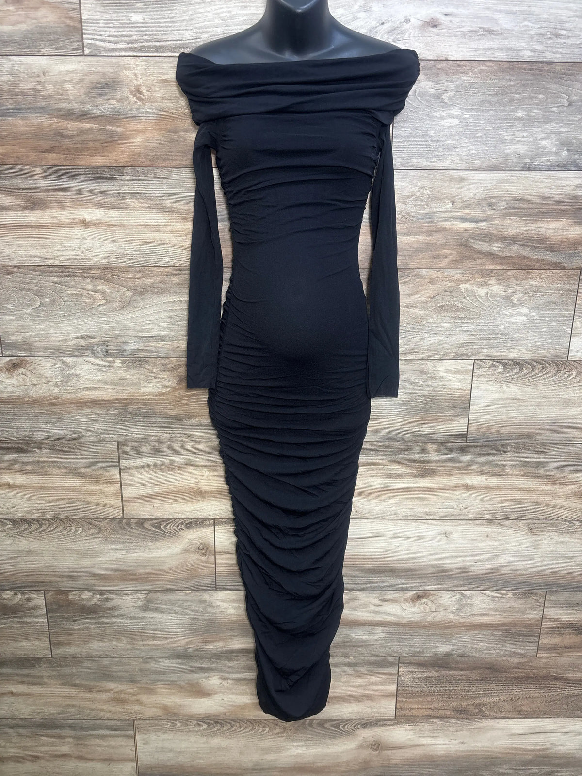 Front view of black Bumpsuit maternity maxi dress in size Small showcasing an off-the-shoulder ruched mesh overlay on a wood-look floor