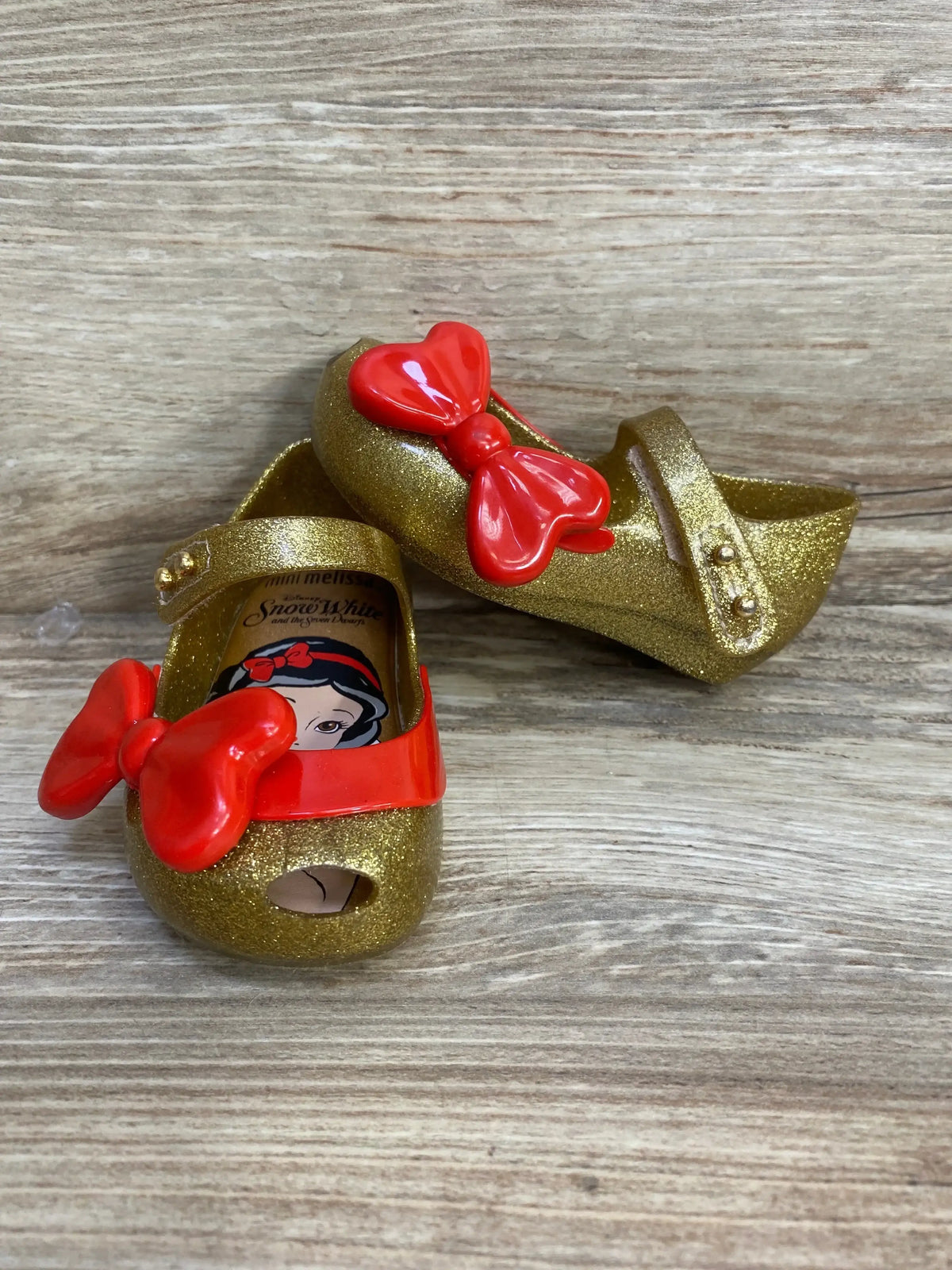 Front and side view of toddler Mini Melissa Ultragirl gold glitter shoes showcasing red bows on a wood background