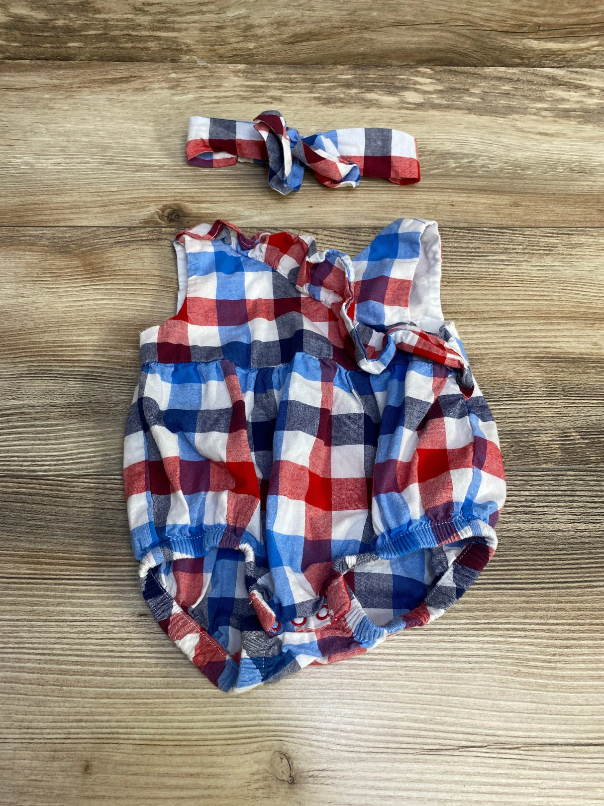 Flat-lay of newborn red, white and blue plaid romper with ruffle shoulder and matching headband on wood tabletop