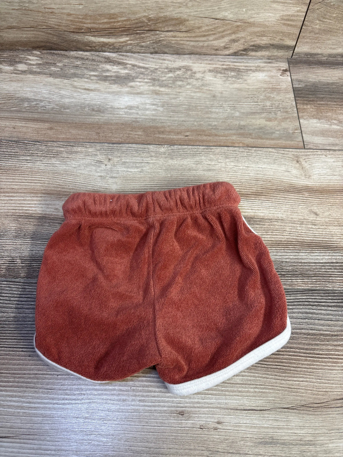 Front view of Little Co. Terry Dolphin Shorts in Adobe Cup showcasing white-trimmed hems and elastic waistband on wood