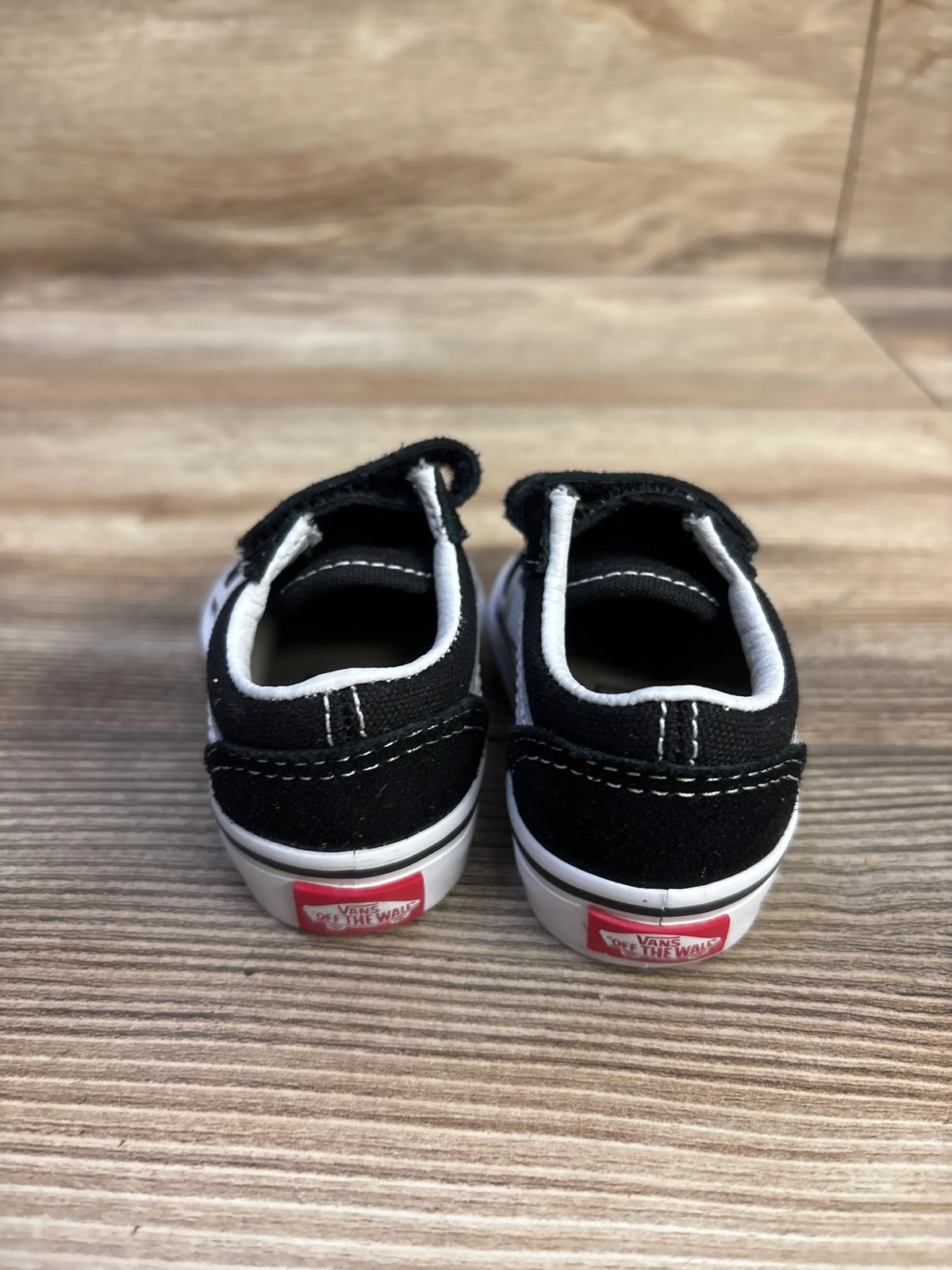 Rear view of black Vans Old Skool V sneakers in size 2C showcasing white trim and red heel labels on a wood floor