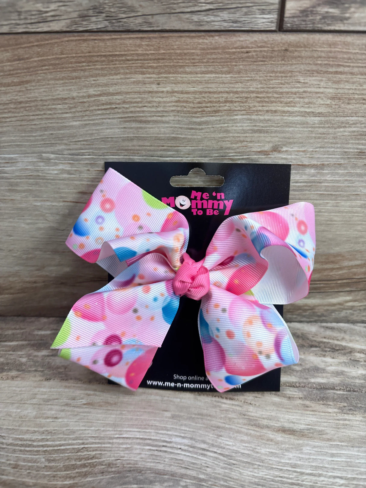 Front view of pink balloon hair bow with multicolored bubble print and knotted center on black card over wood background