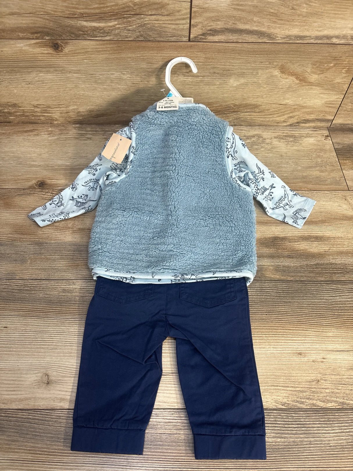 Infant 3-piece outfit with long-sleeve dinosaur print shirt, blue faux fur vest, and navy pants on wood floor