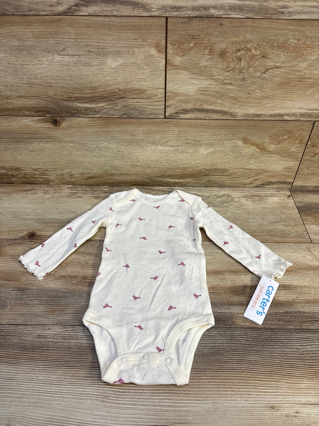 Flat lay of ivory ribbed long-sleeve baby bodysuit (12M) showing small pink bird print and three snap closures on wood background