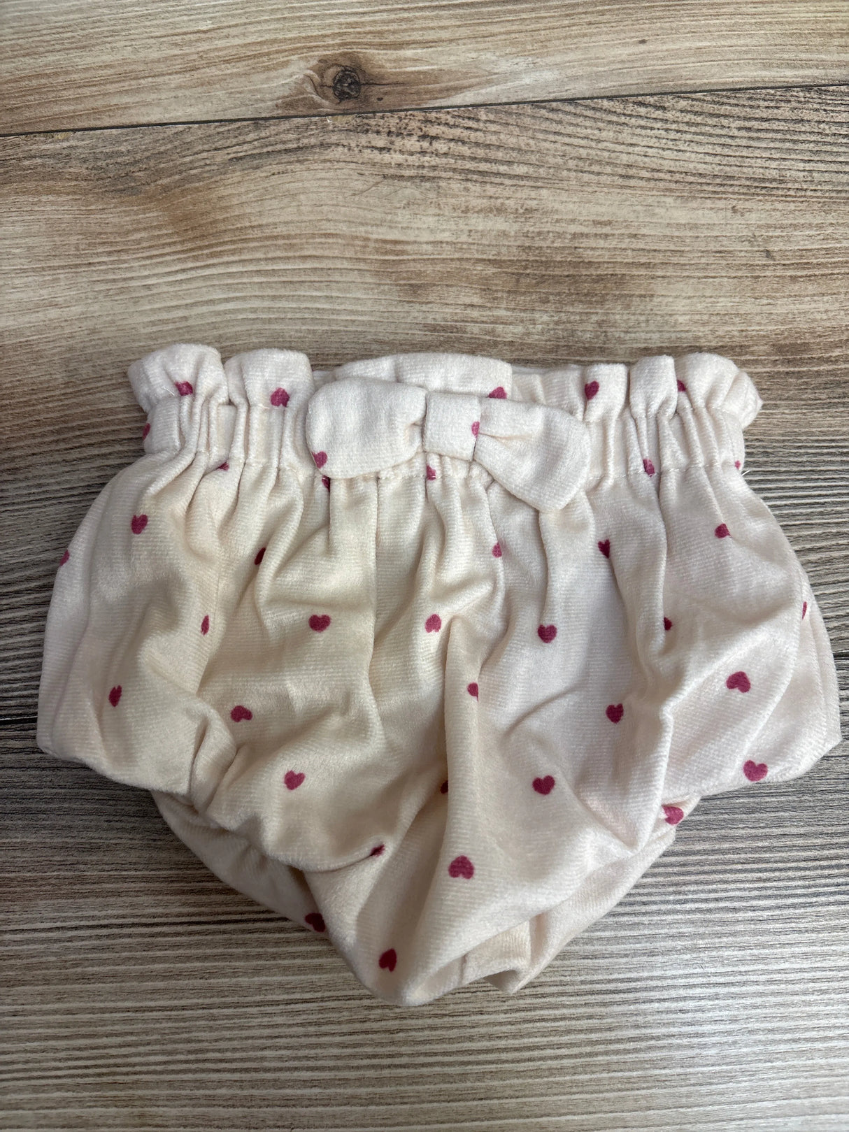 Cream-colored baby bloomers with pink heart print and a fabric bow on wooden background