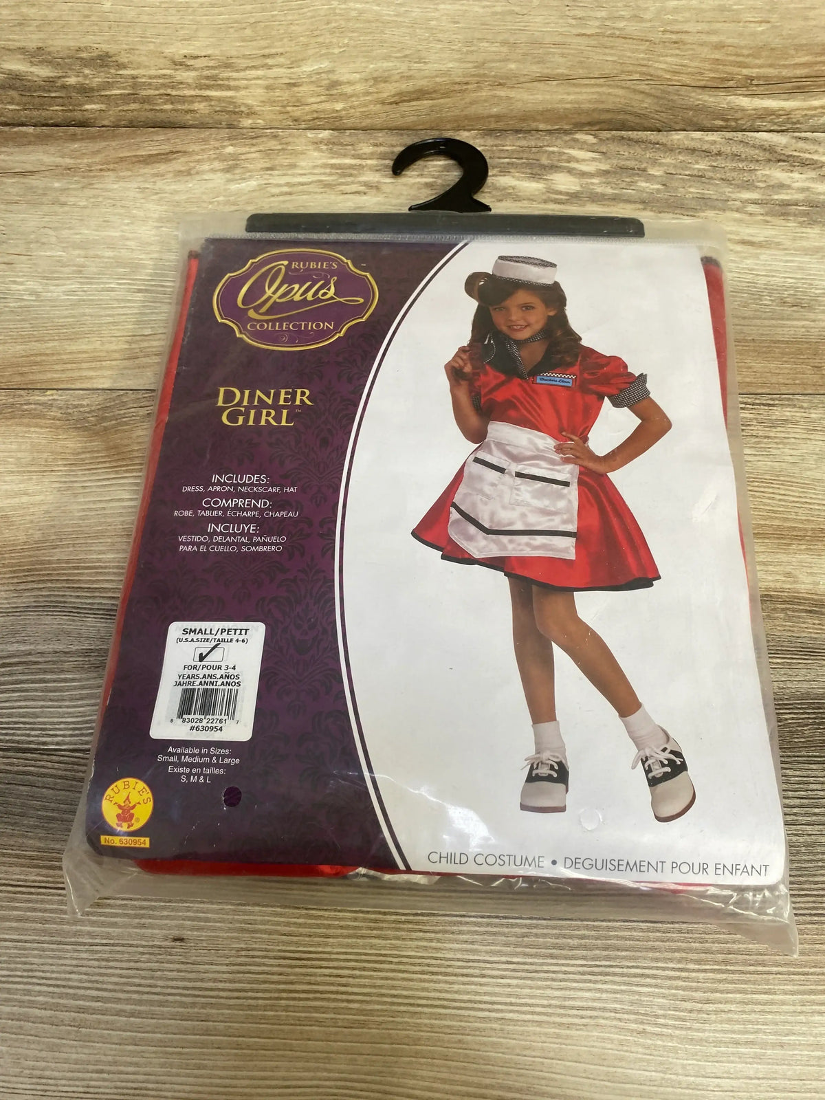 Front view of Rubies Opus Collection Diner Girl costume in red, showing dress, apron, neck scarf, and hat in package on a wood surface