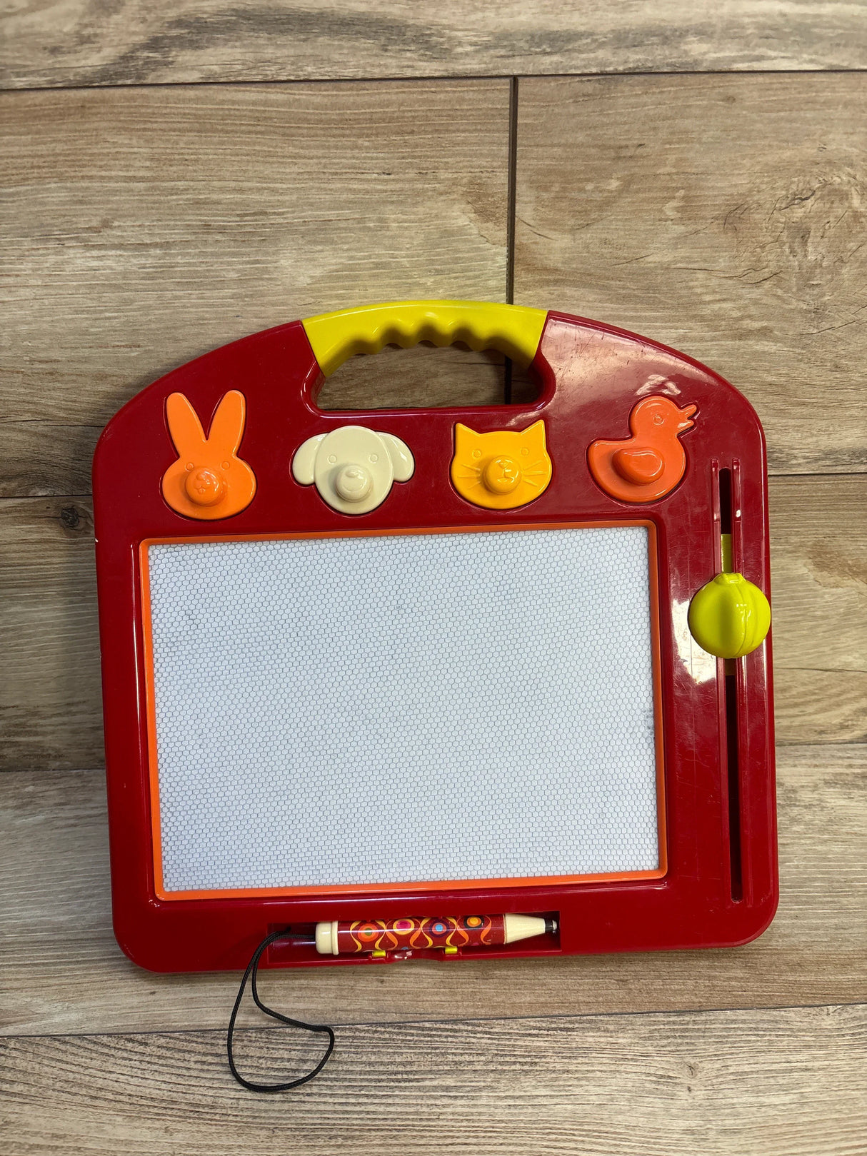 Front view of B. Toys LapTrec magnetic board with animal stamp knobs, stylus, and yellow erase slider on wood floor