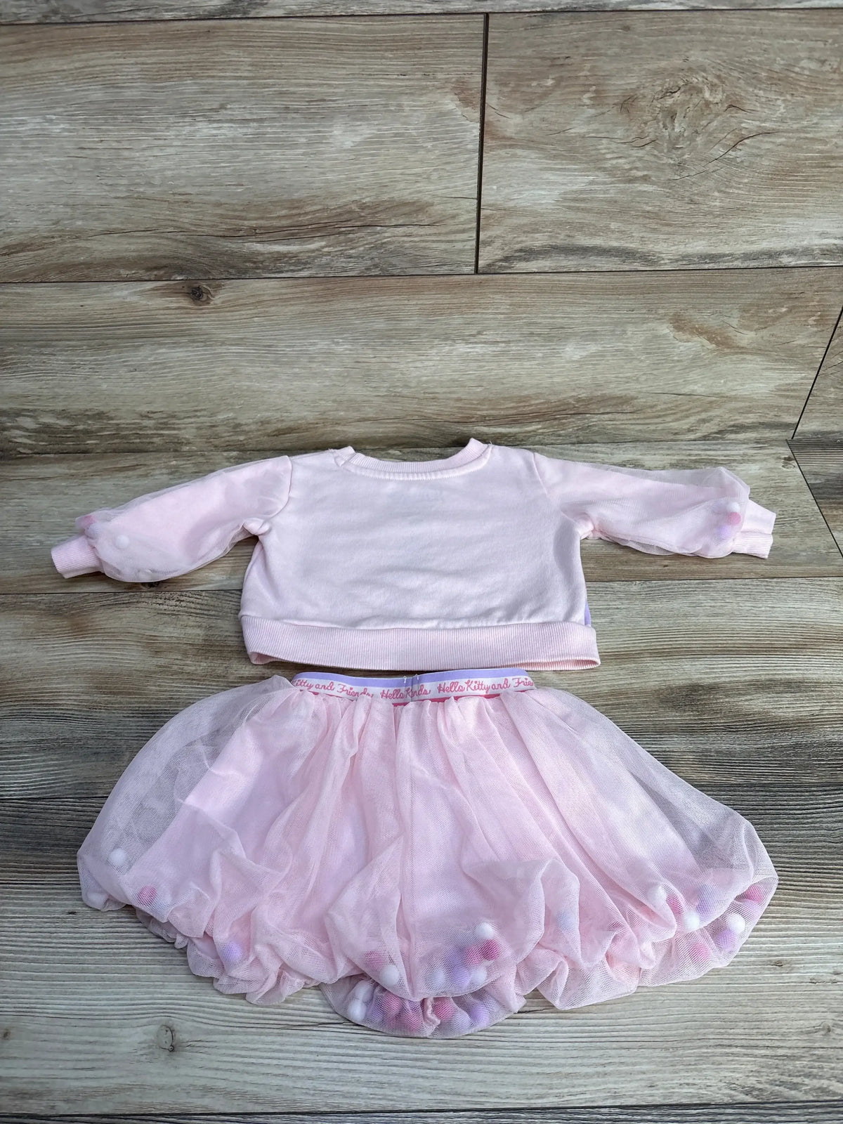 Flat lay of pink Hello Kitty sweatshirt and tulle skirt, 12 months, showing sheer puff sleeves and pom-pom tulle hem on wood floor
