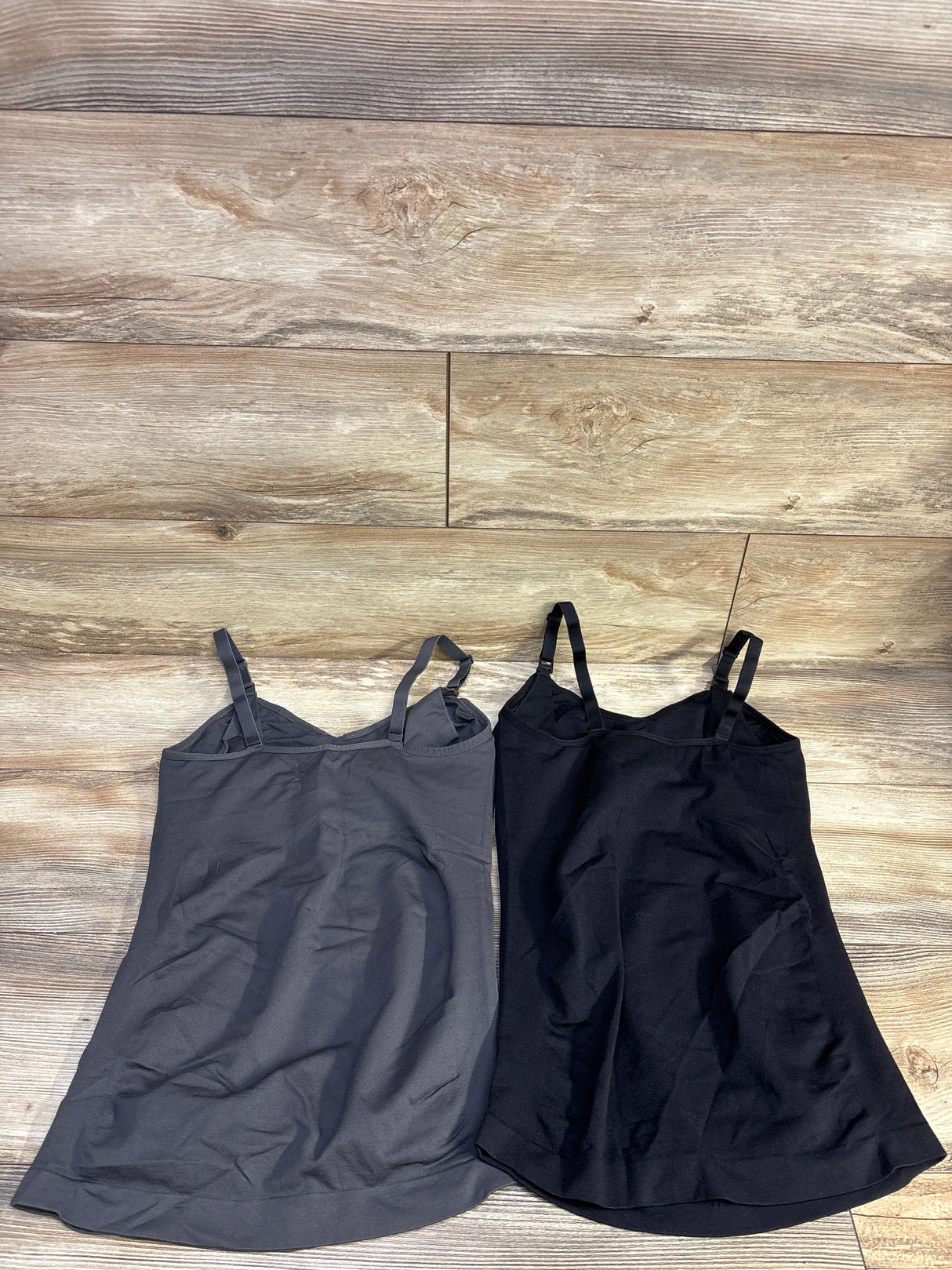 Flat lay of two nursing tank tops (grey and black) with adjustable shoulder straps and inner shelf on wood floor background