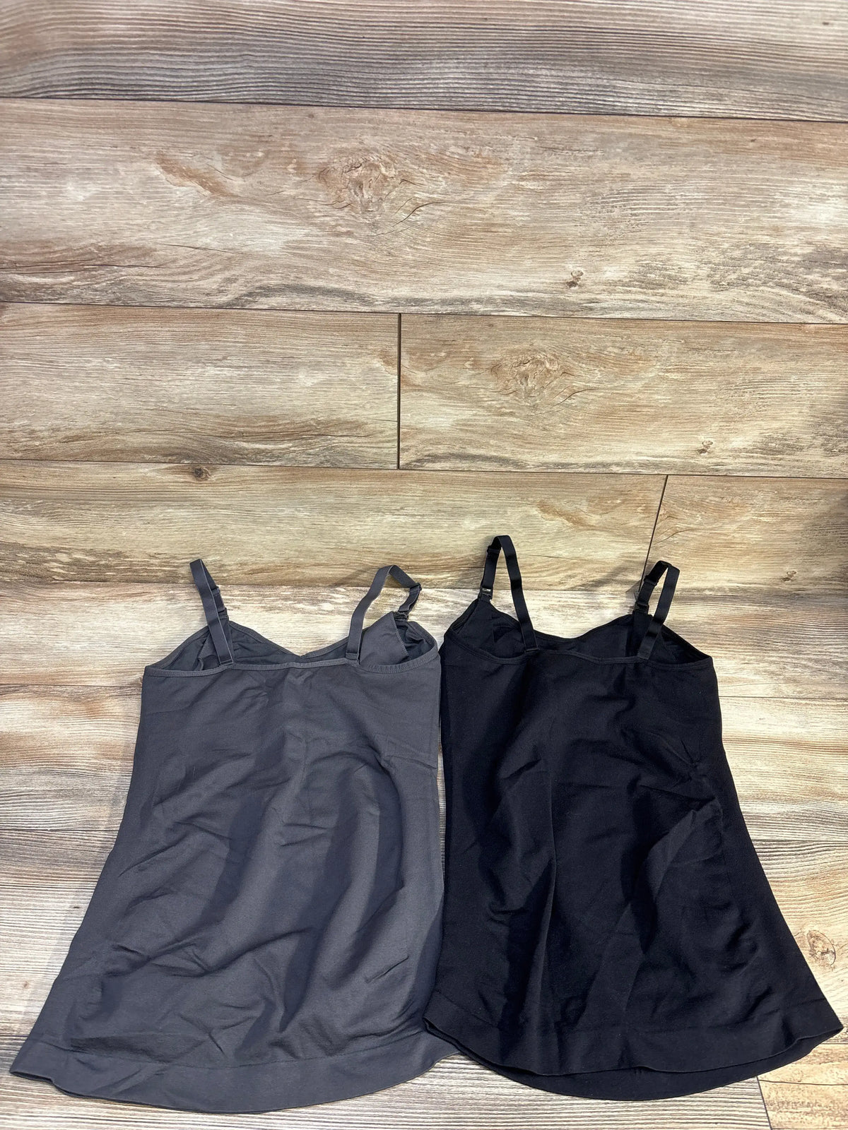 Flat lay of two nursing tank tops (grey and black) with adjustable shoulder straps and inner shelf on wood floor background