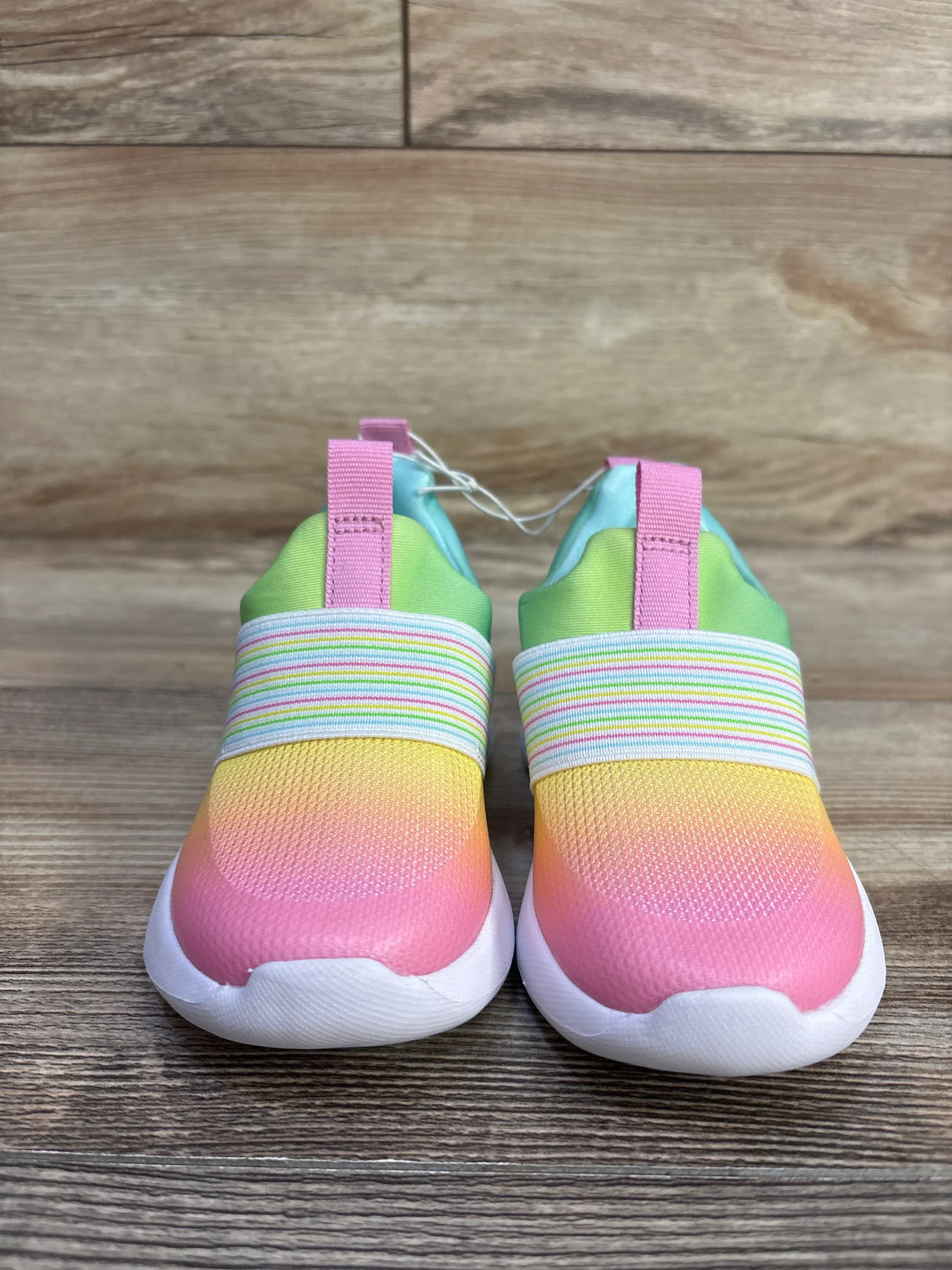 Front view of Cat & Jack toddler slip-on sneakers showing rainbow gradient toe, striped elastic band and pink heel loops on wood floor