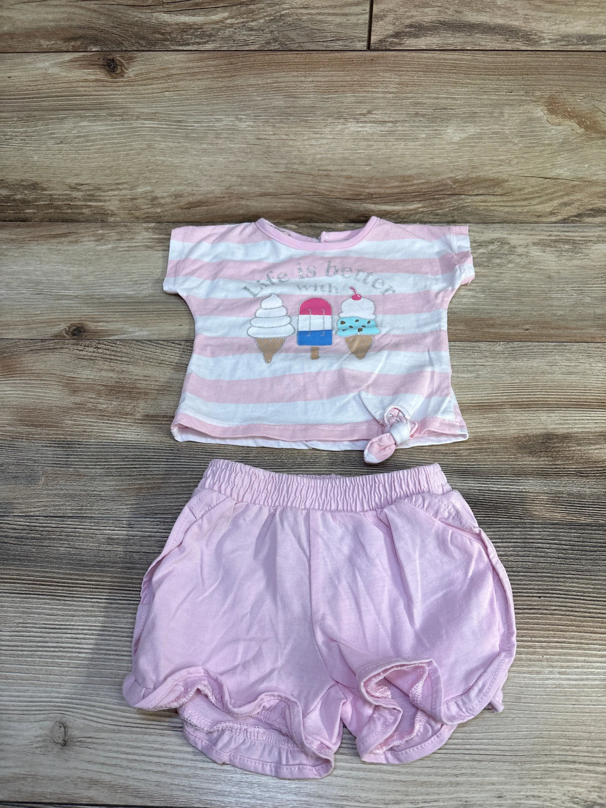Flat lay of pink baby 2pc set: tee with ice cream graphic saying Life is better with ice cream, knotted hem, shorts on wood
