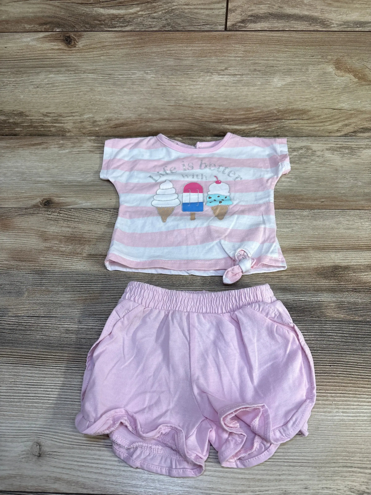 Flat lay of pink baby 2pc set: tee with ice cream graphic saying Life is better with ice cream, knotted hem, shorts on wood