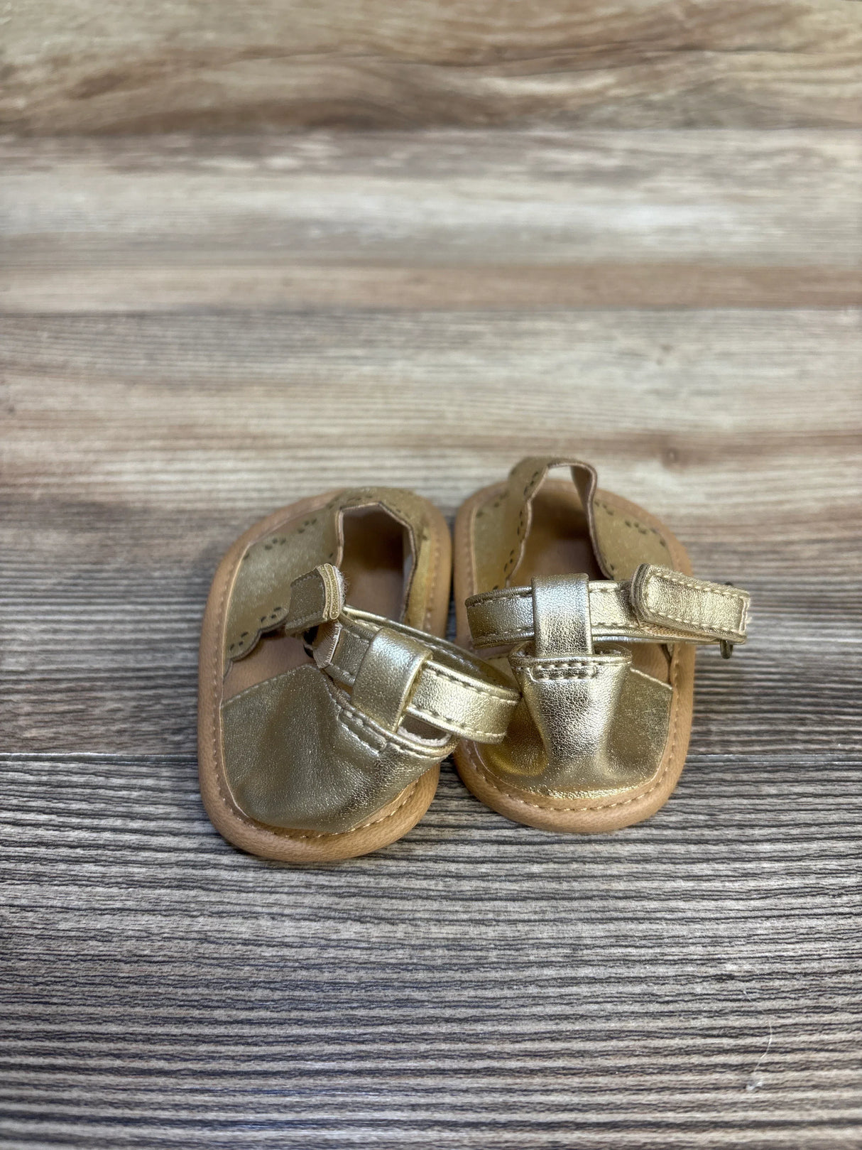 Front view of Old Navy gold faux-leather baby sandals size 1/2C 0-3M with scallop trim and Velcro ankle straps on wood