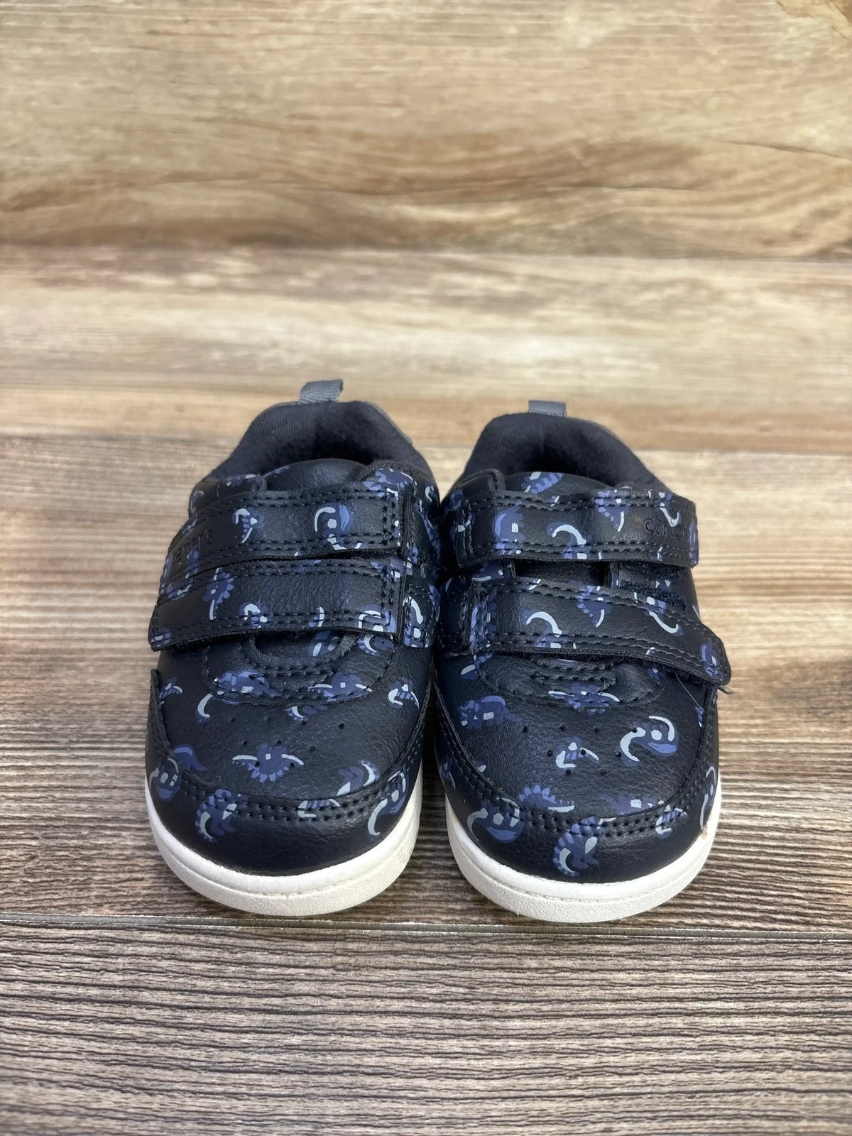 Front view of navy Carter’s Neo Dino First Walker sneakers with two hook-and-loop straps and pull tabs on wood background