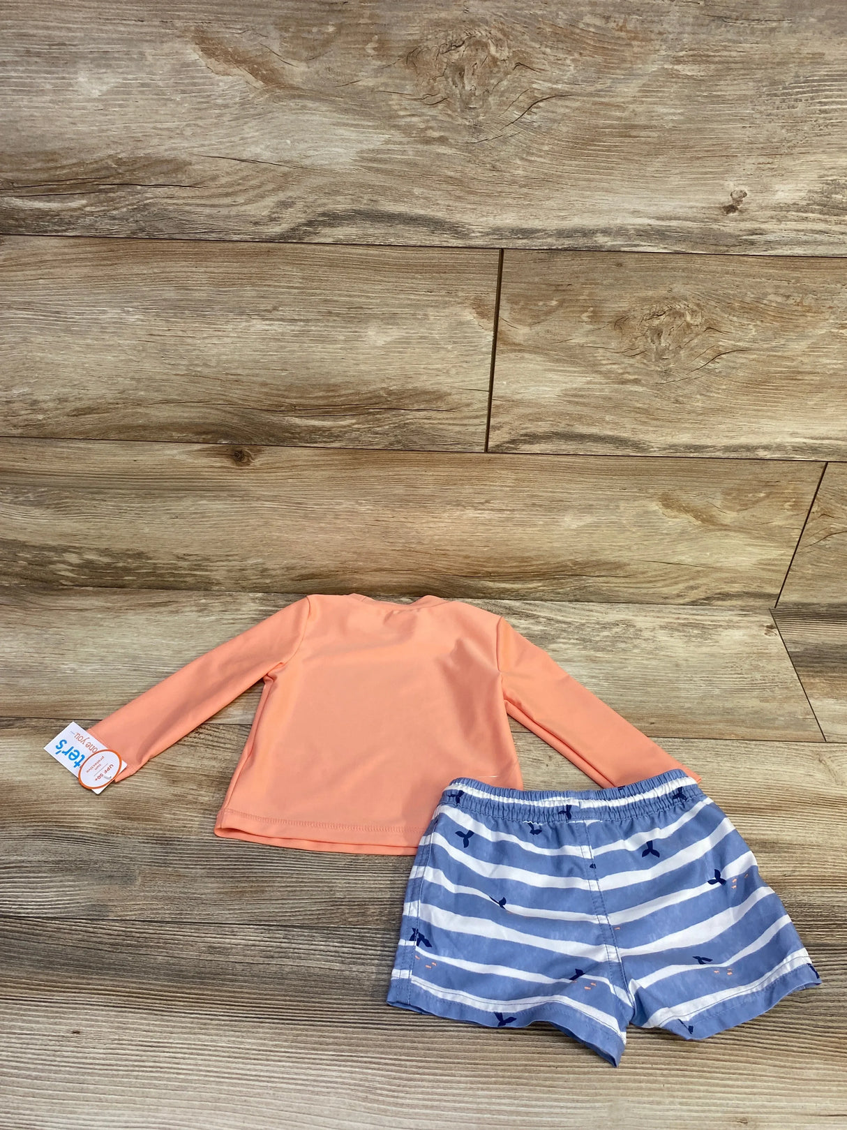 Flat lay of baby 2-piece rash guard set: coral long-sleeve top and blue striped whale swim shorts on wood background