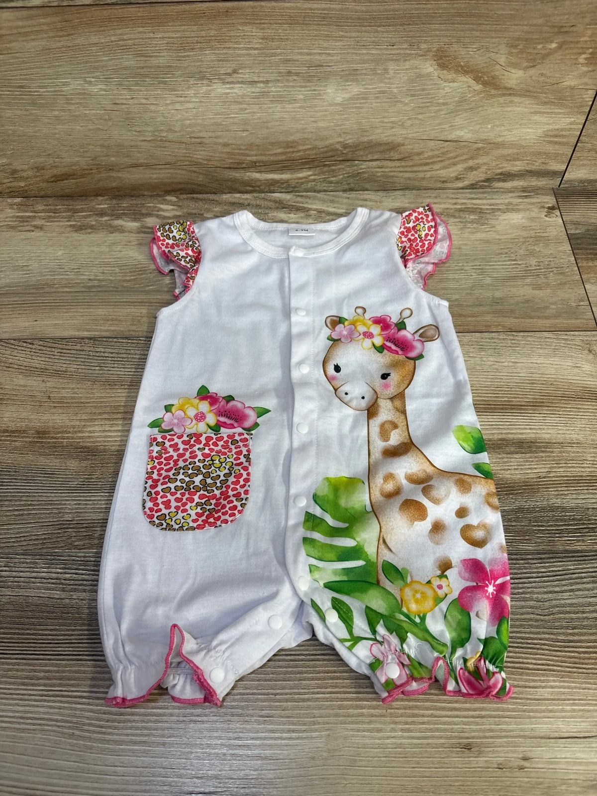 Flat lay of white baby romper showing giraffe and tropical floral print, front snap closures, ruffled cap sleeves and pocket on wood background