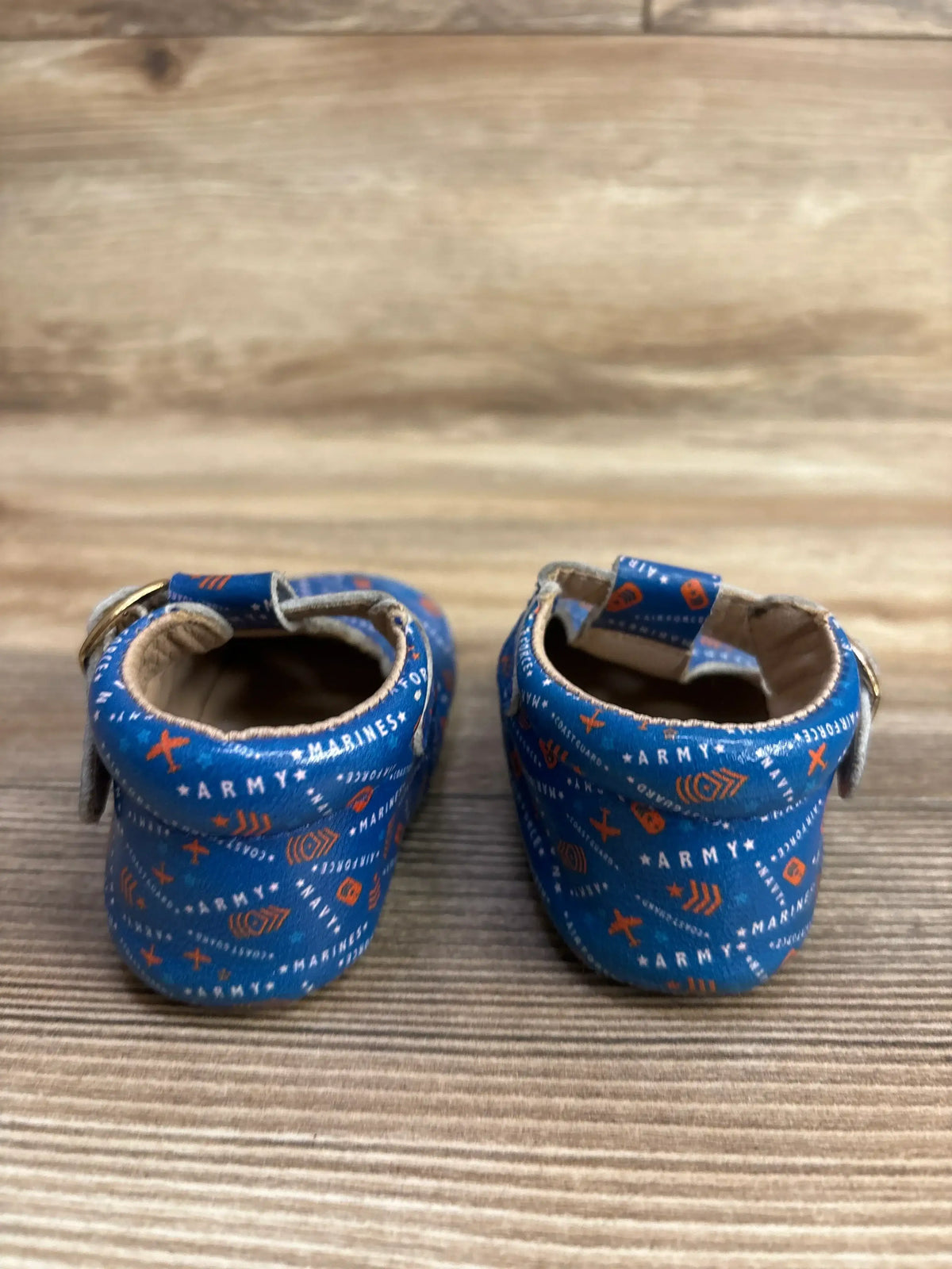 Rear view of Monkey Feet T-strap soft-sole baby shoes showcasing blue military branch print and buckle strap on wooden background