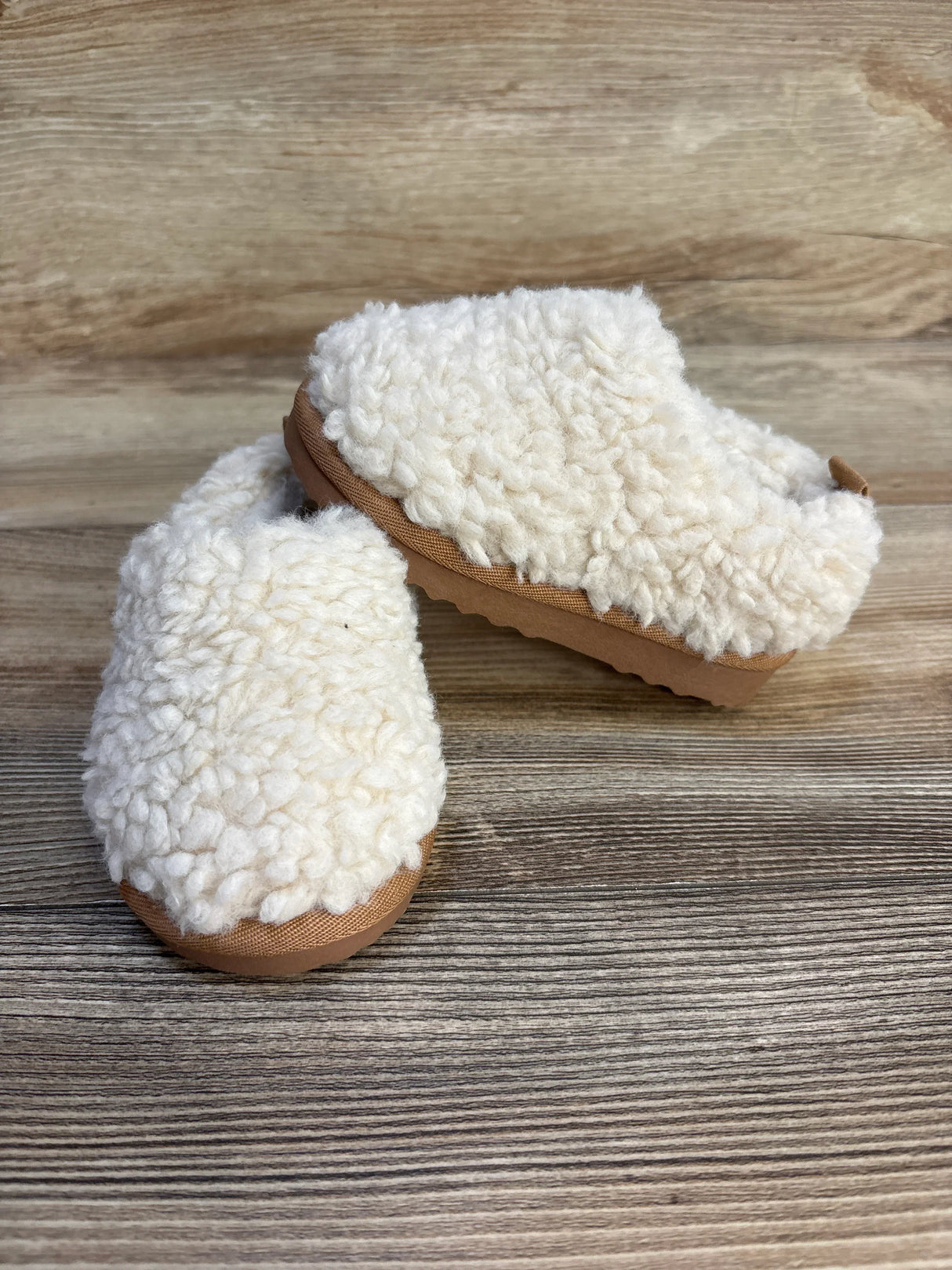Pair of cozy cream toddler sherpa slippers with tan soles on wood background