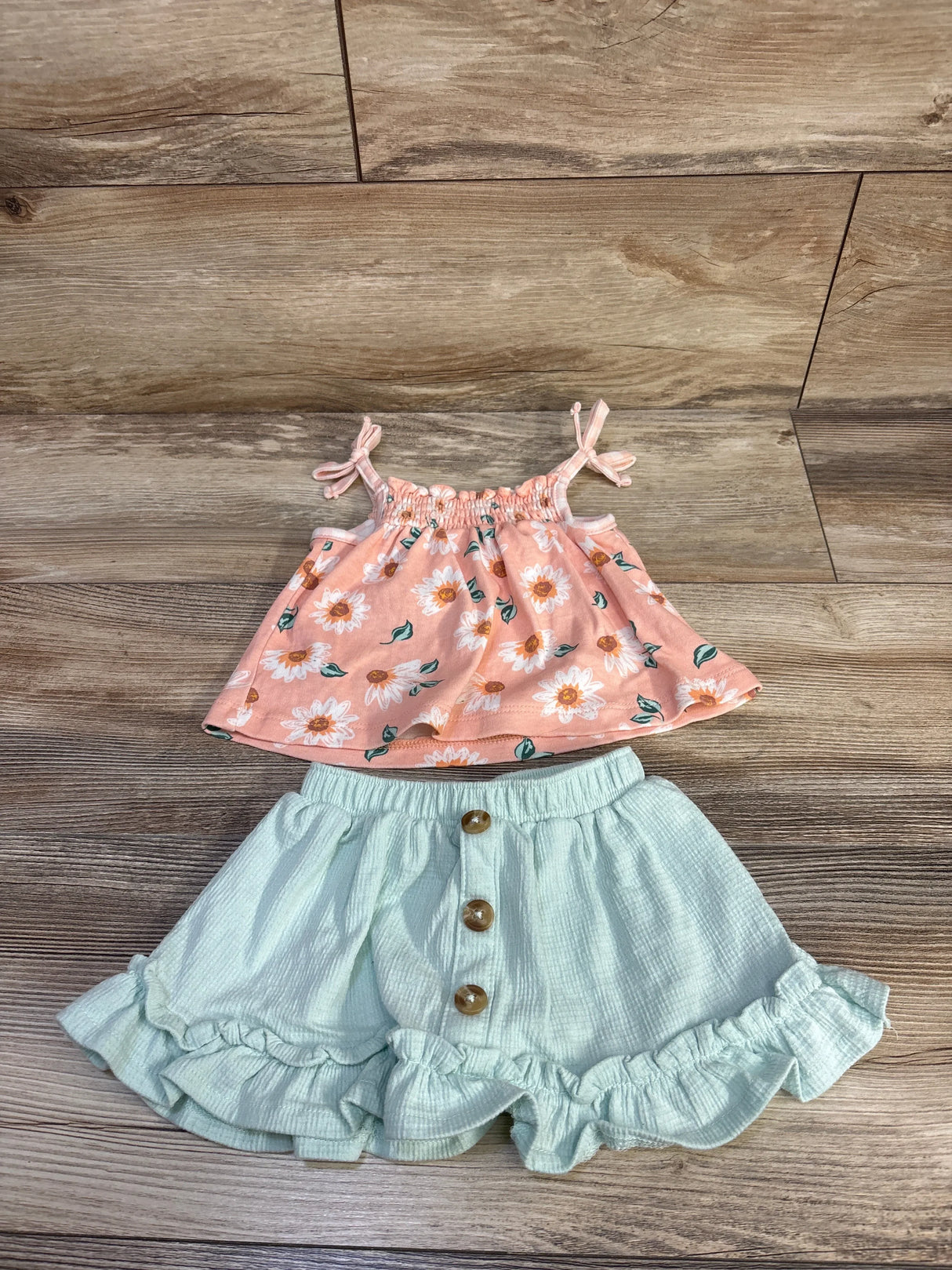 Flat lay of pink floral tank top with tie straps and mint green ruffled shorts with wooden button front on light wood floor