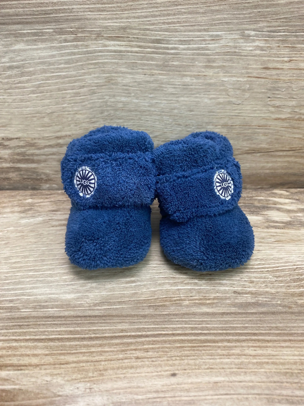 Front view of UGG Baby Bixbee boot pair in blue showing plush terry fleece material and UGG logo strap on wooden background size 0C/1C