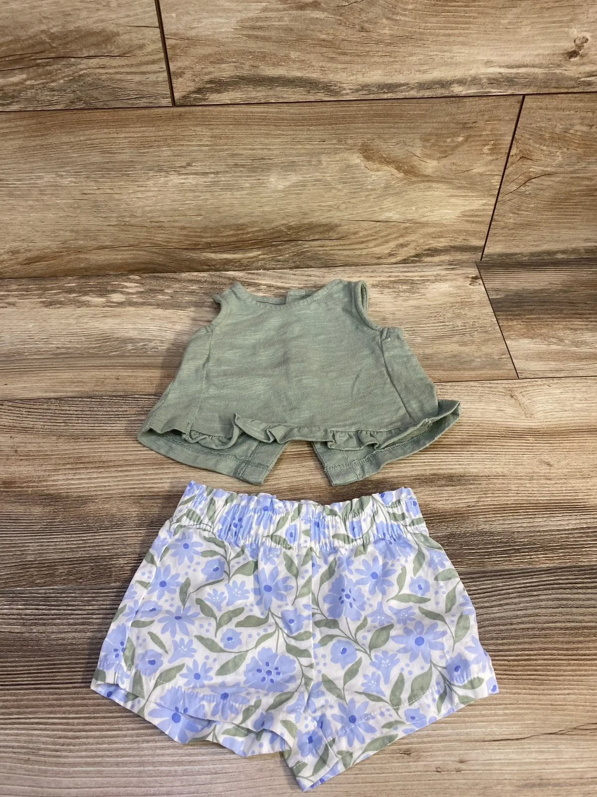 Flat lay of Just One You 2pc set size 3m showcasing sage green tank top and white shorts with blue floral print on wood plank floor