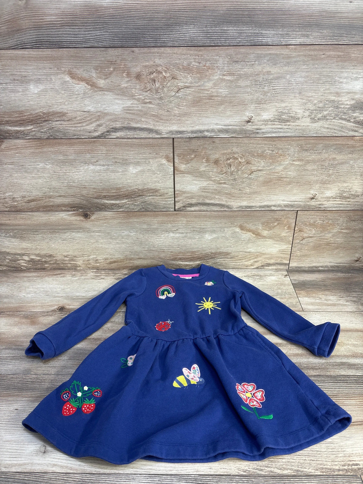 Front view of a navy Mini Boden embroidered sweatshirt dress showcasing colorful embroidered motifs on a wood background