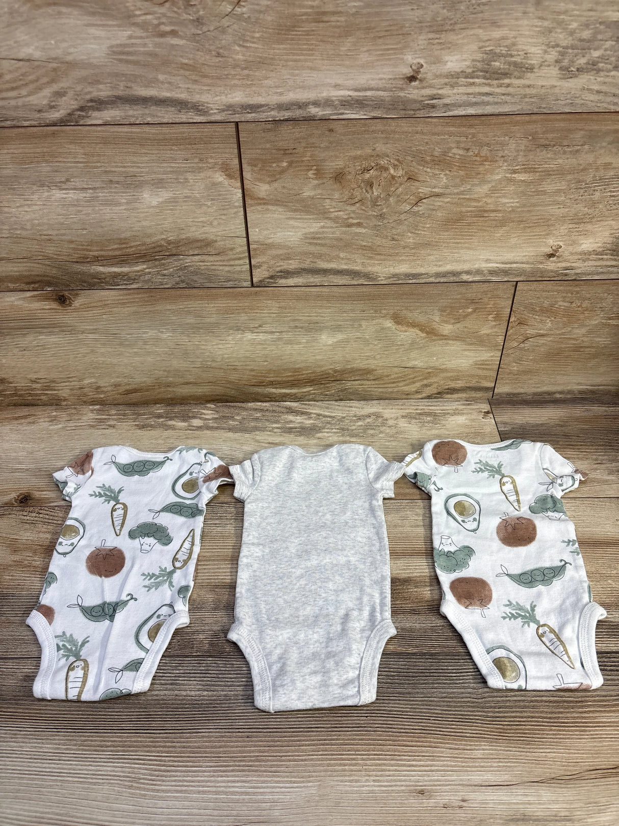 Flat lay of three newborn bodysuits showing vegetable print on two and heather gray bodysuit in center on wood floor
