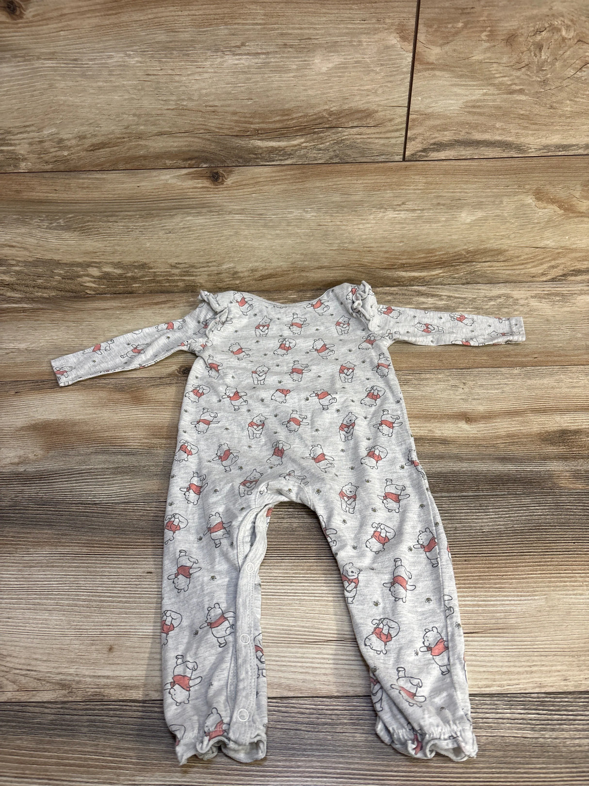 Flat lay of grey Winnie the Pooh coverall size 18M showing all-over print, leg snaps and ruffle shoulders on wood floor