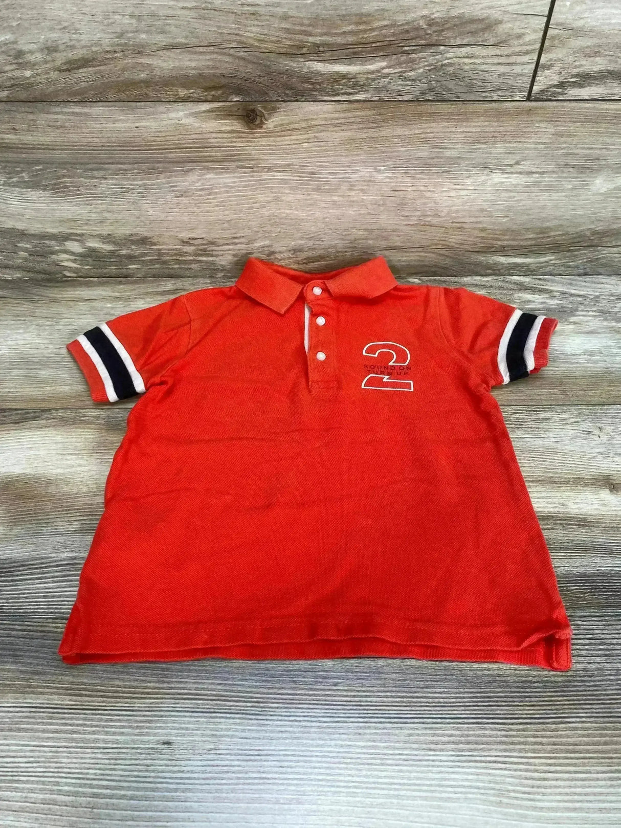 Front view of red Mayoral toddler polo shirt showing three-button placket, black and white striped sleeve cuffs and number 2 logo on wood background
