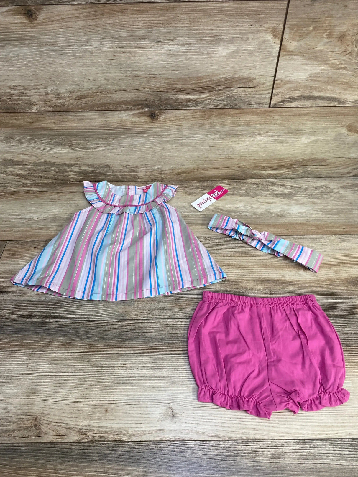 Flat lay of Penelope Mack 3pc baby outfit with striped ruffle top, matching headband, and pink elastic bloomers on wood