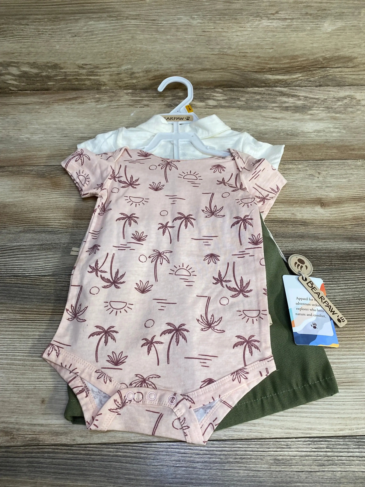 Flat lay of 3-piece Bear Paw Paradise short set showing pink palm-print bodysuit, white top, and olive shorts on wood background