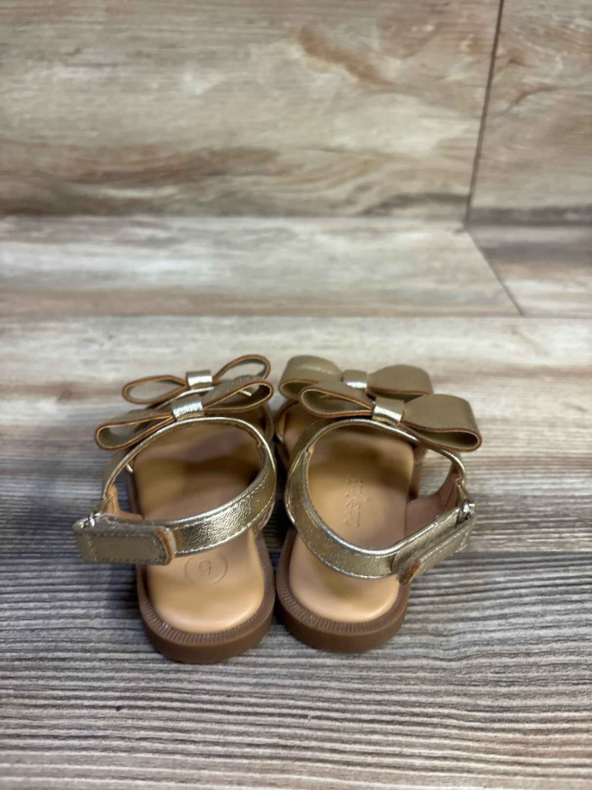 Back view of Cat & Jack bow sandals in metallic gold showing double bows, Velcro straps, and brown soles on wood floor